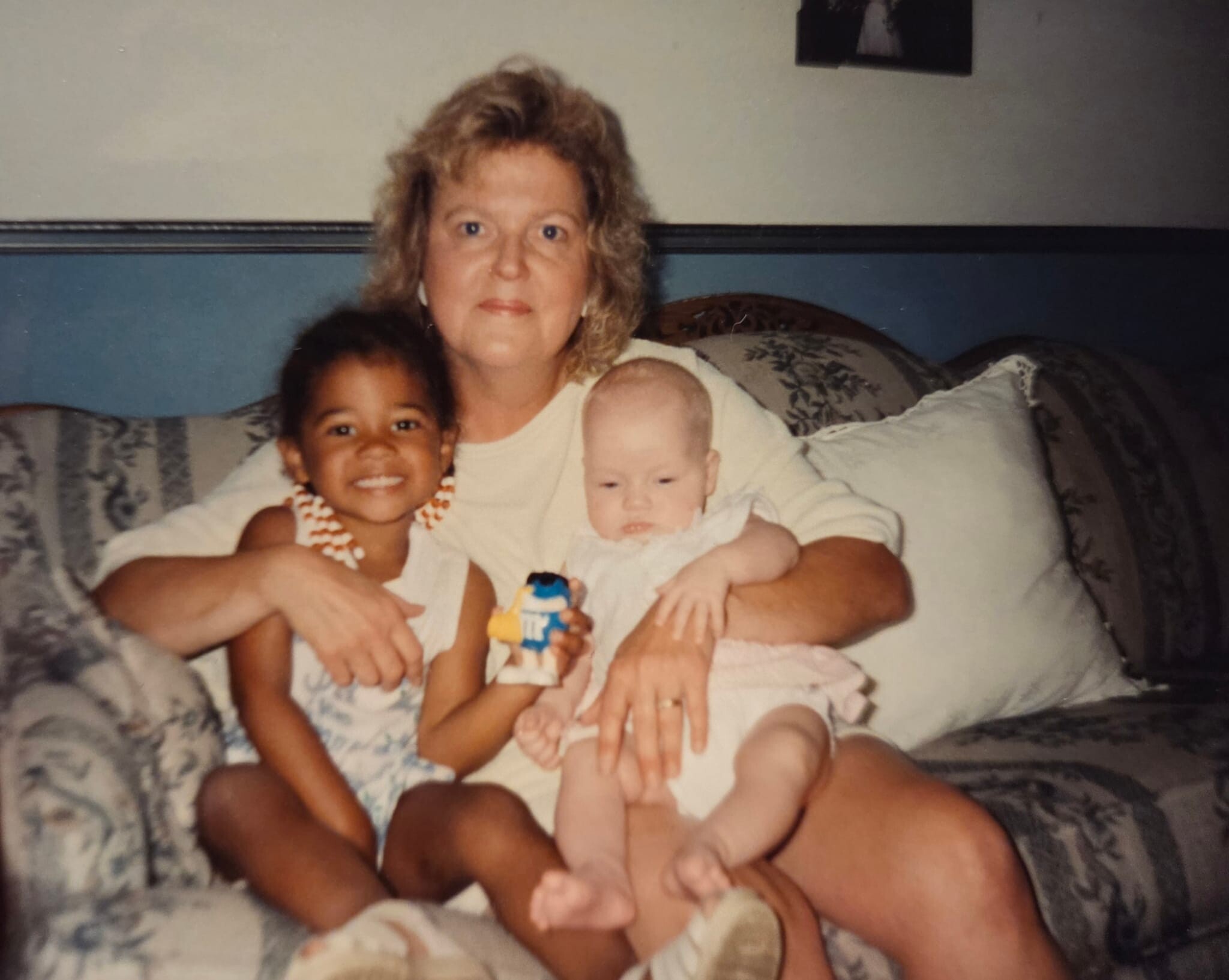 Mother sitting on a patterned couch, holding a baby and a toddler nearby, indoors in a home setting.