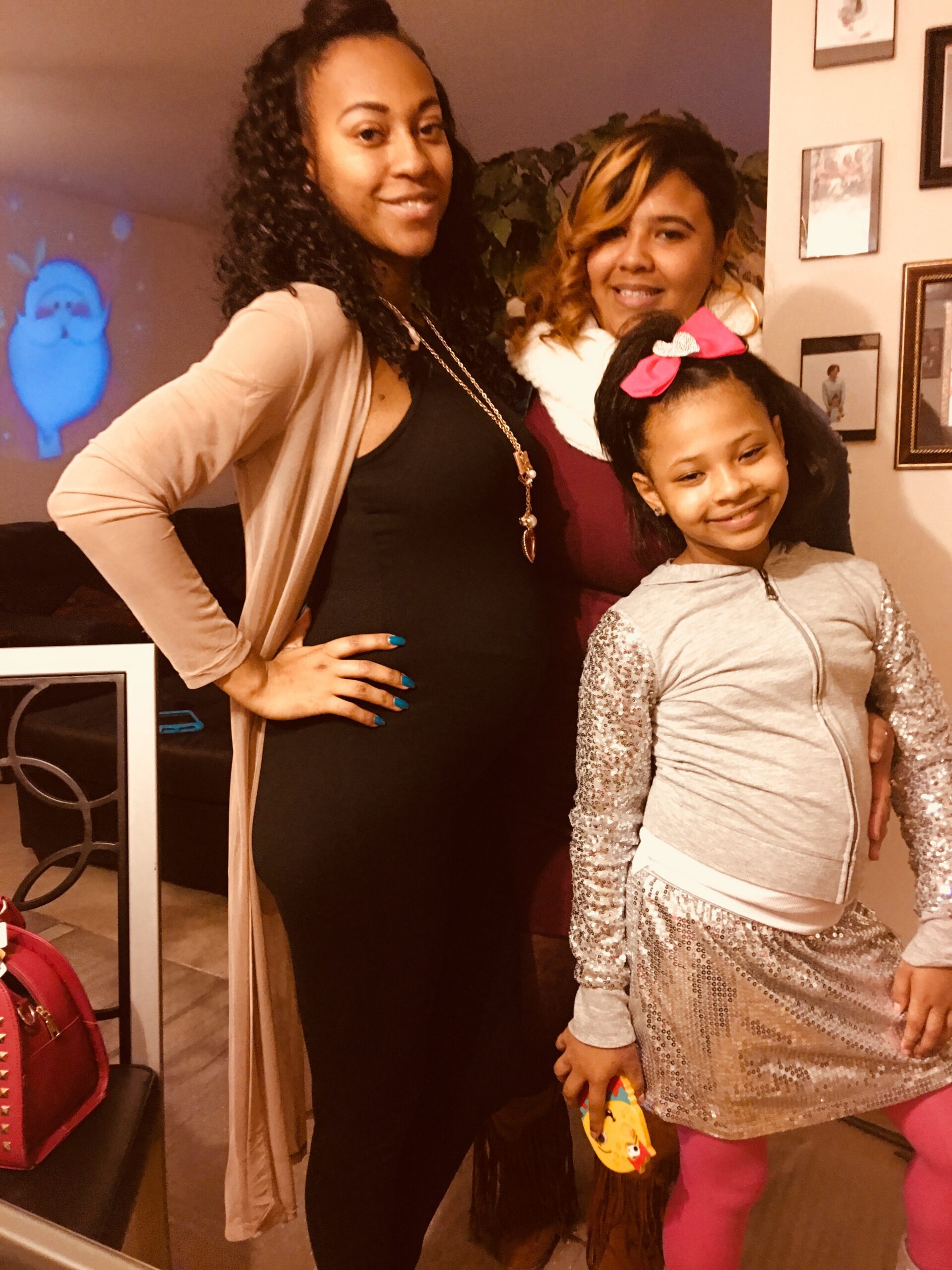 Three people posing indoors: two women and a girl in a sparkly outfit with a pink bow, all smiling at the camera.