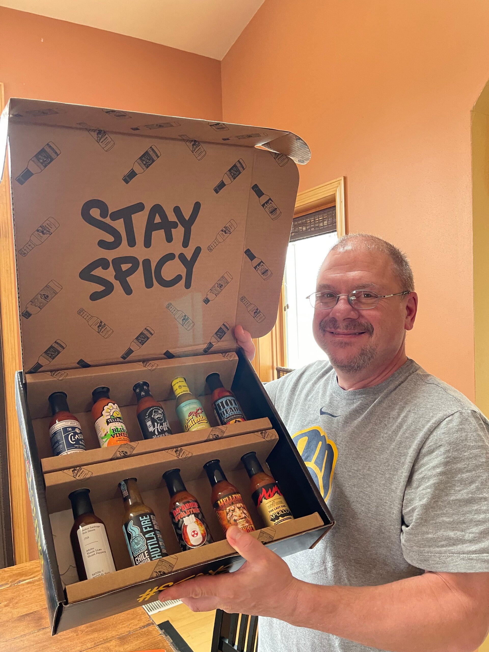 Smiling man in a gray shirt holds an open gift box of assorted craft beer bottles, with 'STAY SPICY' printed on the box.
