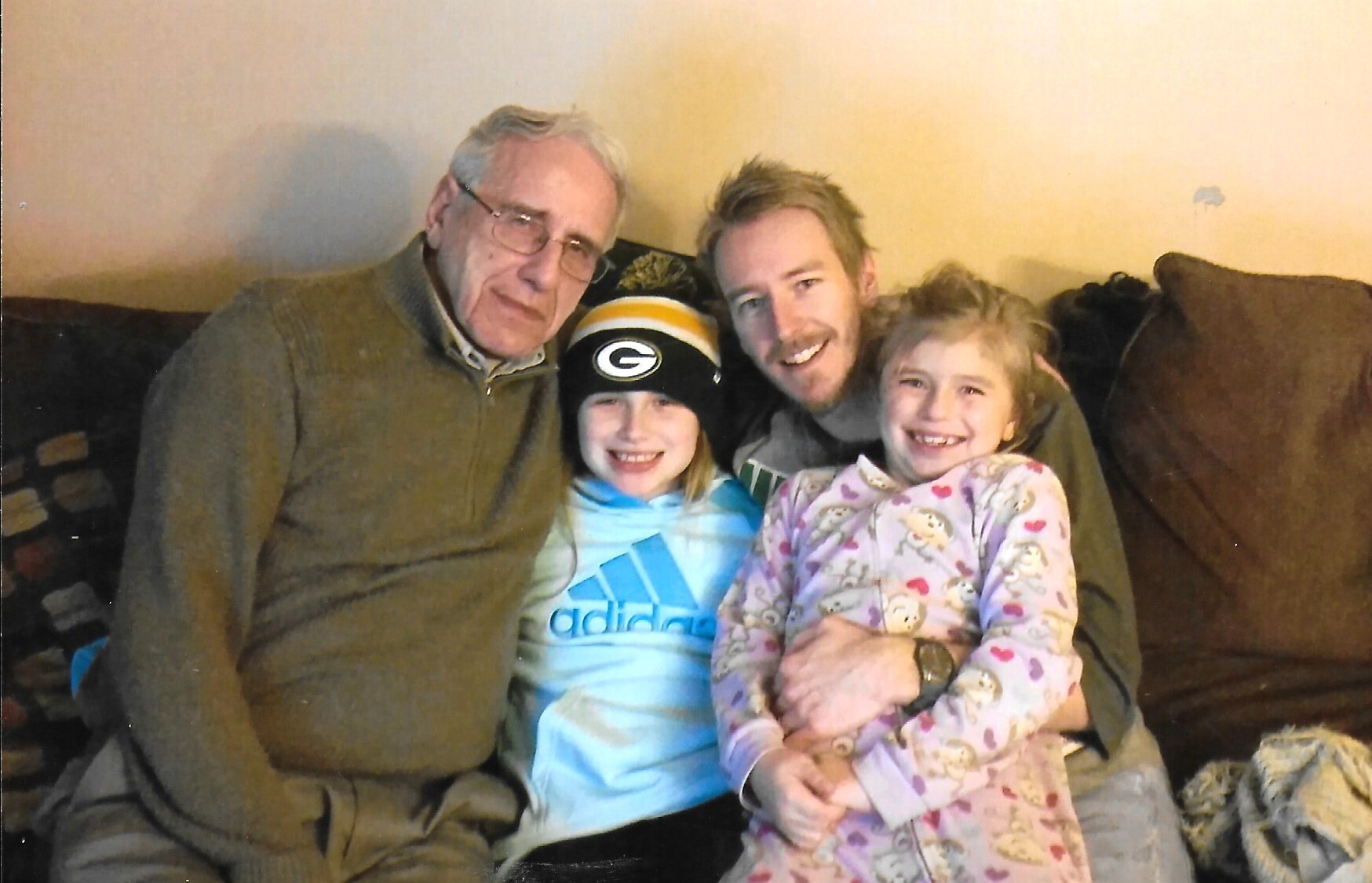 Grandfather, father, and two smiling girls seated together on a couch for a family photo.