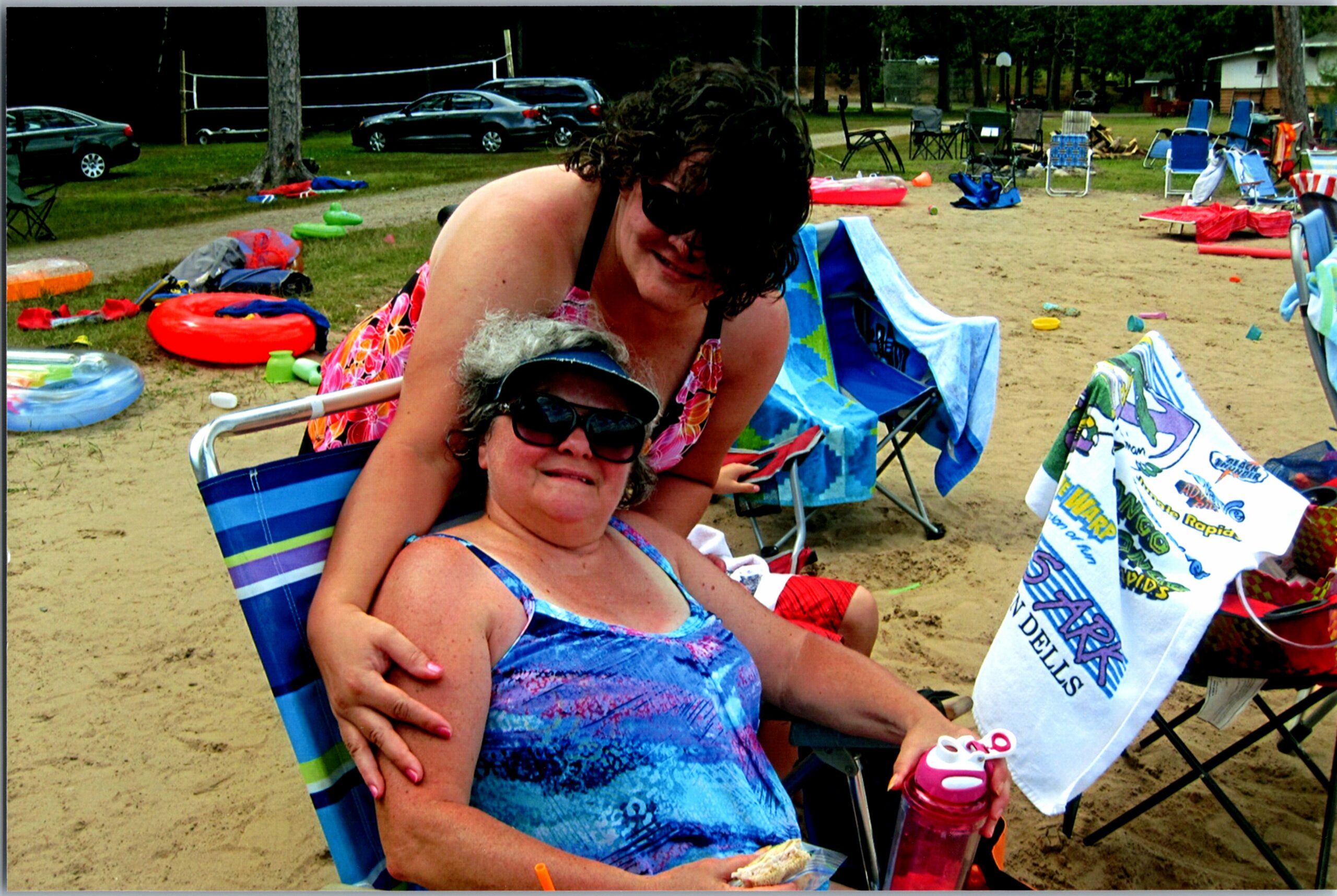 Two women sit close together in folding beach chairs on a sandy lakeside, smiling with towels and gear scattered around during a sunny day.