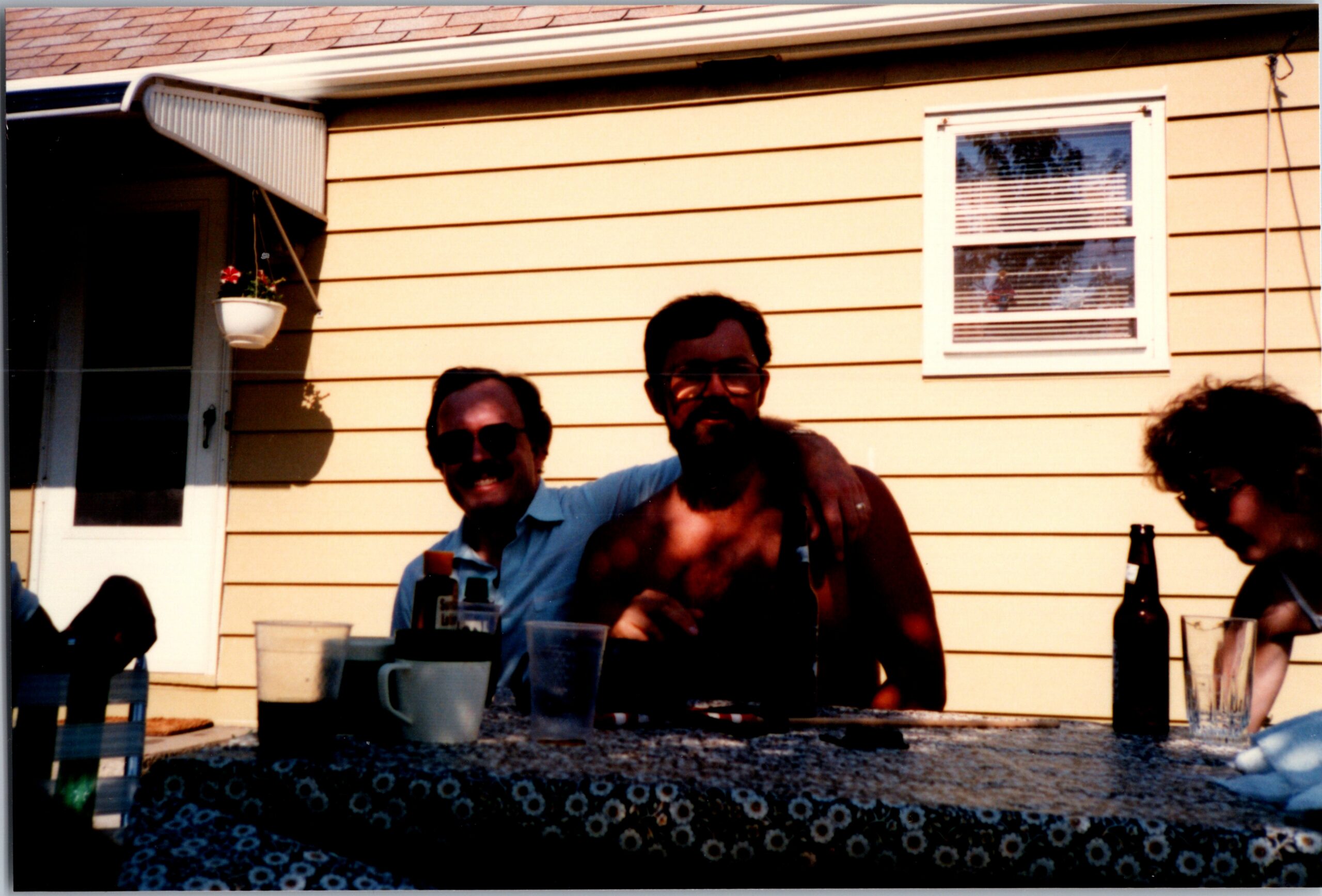 Group of friends at a backyard gathering around a table with cups and bottles, in front of a yellow house with a small window and hanging planter.