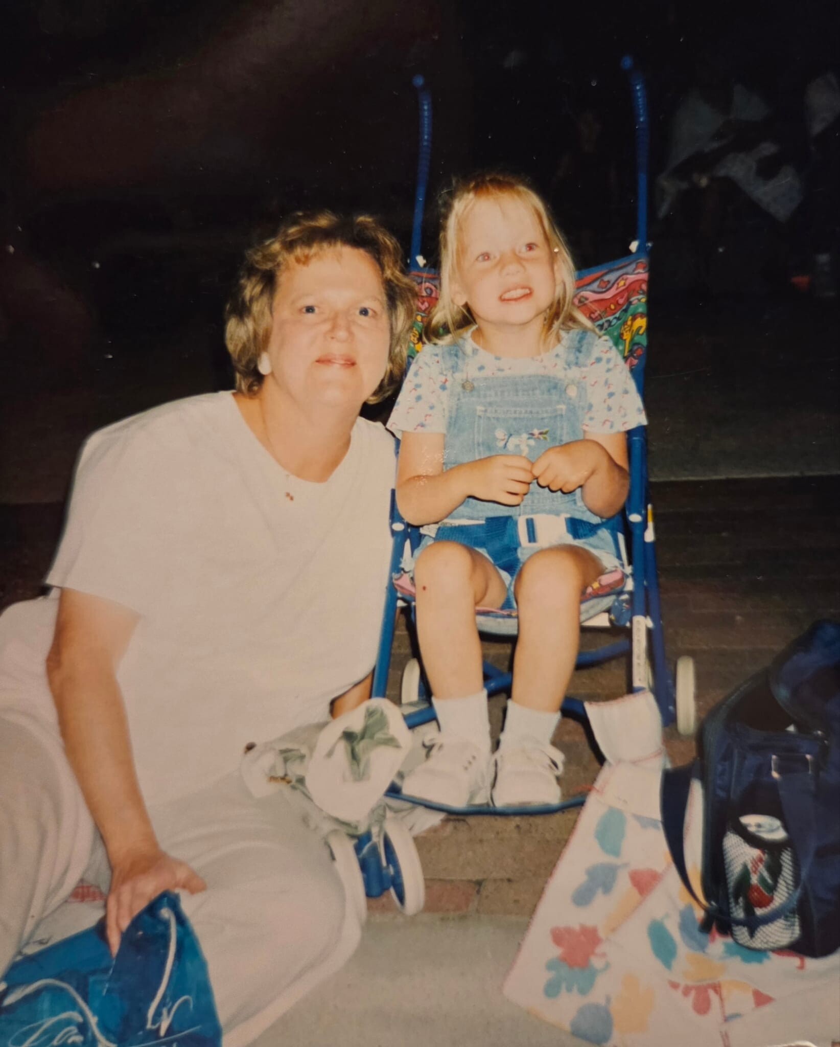 An older woman crouches beside a smiling young girl sitting in a colorful stroller outdoors at night.