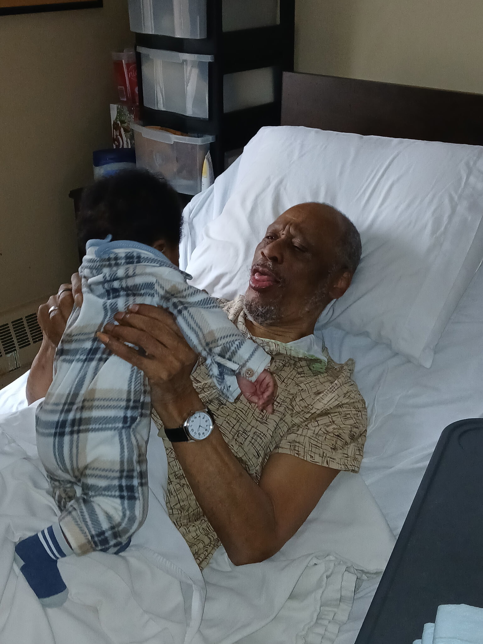 Older man lying in a hospital bed embracing a child who is standing beside him, both facing each other. The man wears a patterned shirt and a wristwatch; a white pillow supports his head. Behind them are hospital storage shelves and medical items.