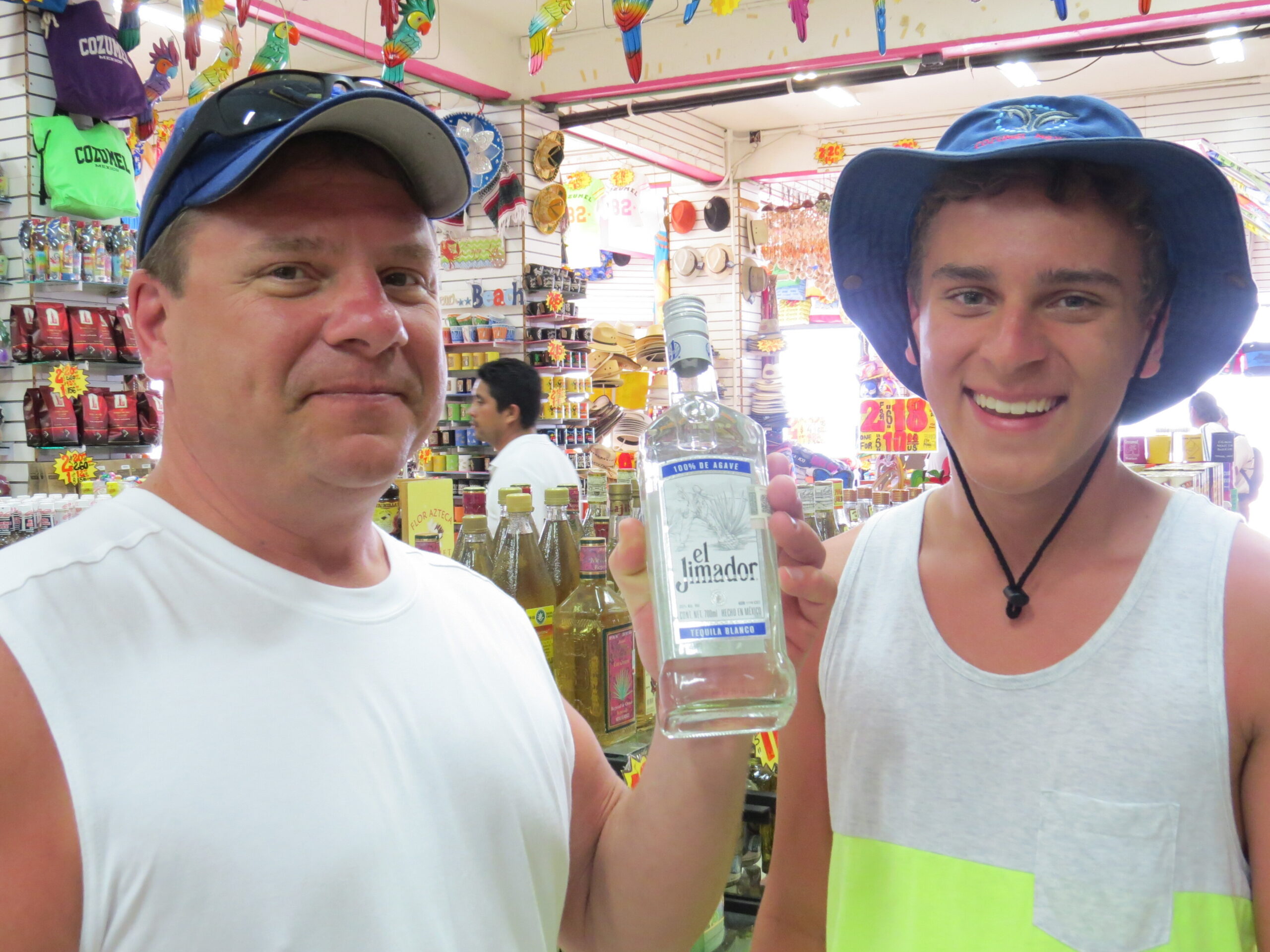Two men in a bright shop posing for a selfie while holding a bottle of El Jimador tequila in the foreground.