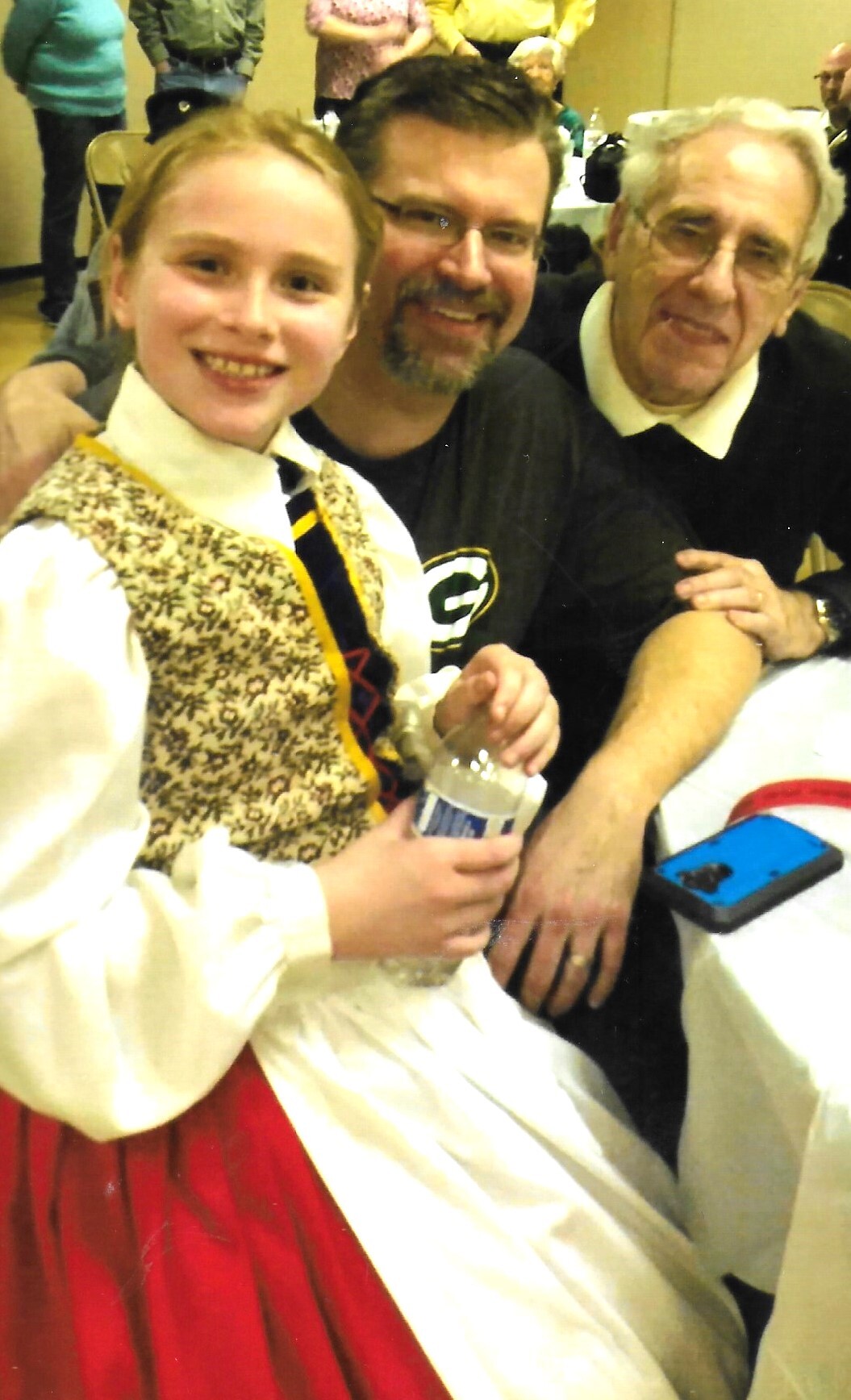 Three people pose for a photo at a social event: a smiling girl in a cream blouse with a patterned vest and red skirt, holding a water bottle, flanked by a man with glasses and a gray-haired man in a dark sweater.