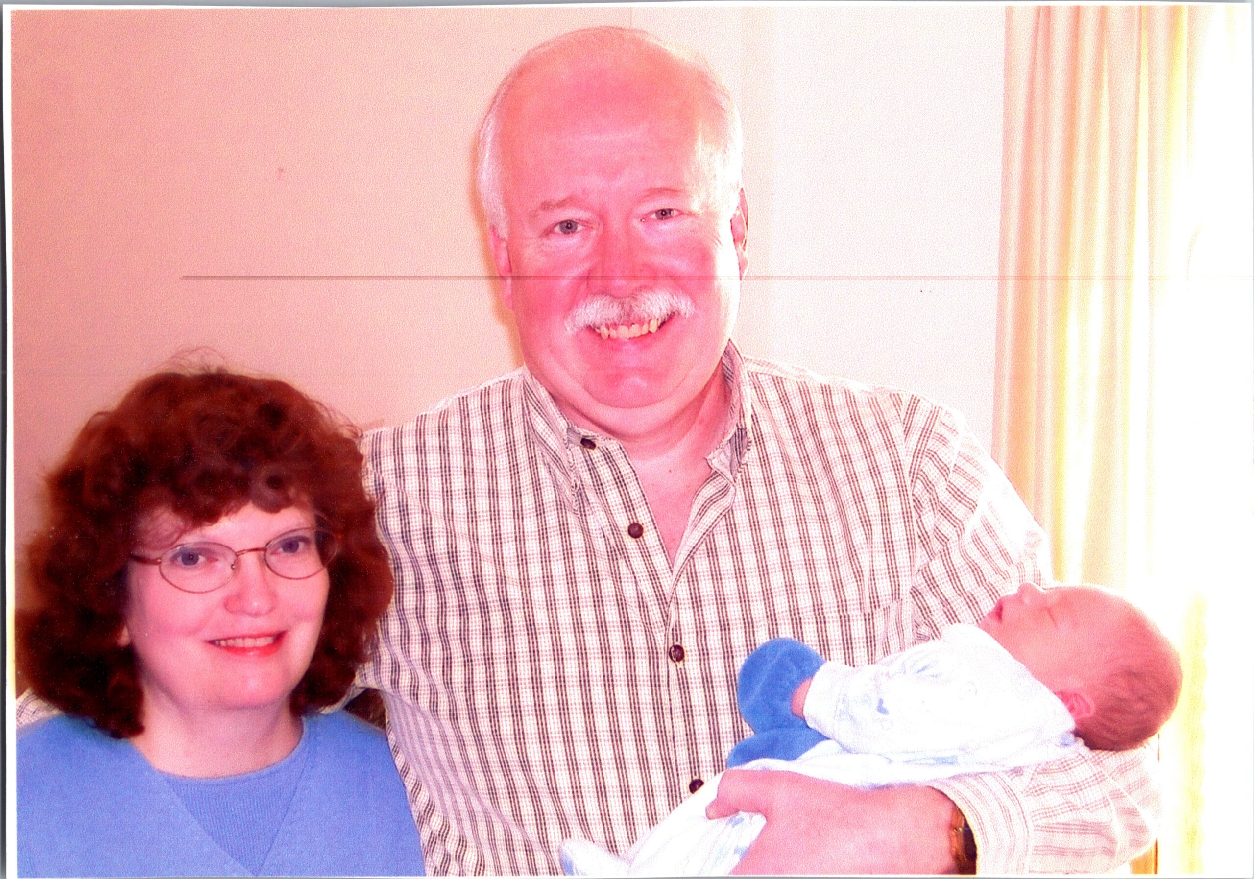 Smiling older man in a plaid shirt holding a baby, with a woman in a blue top beside them in a home setting.