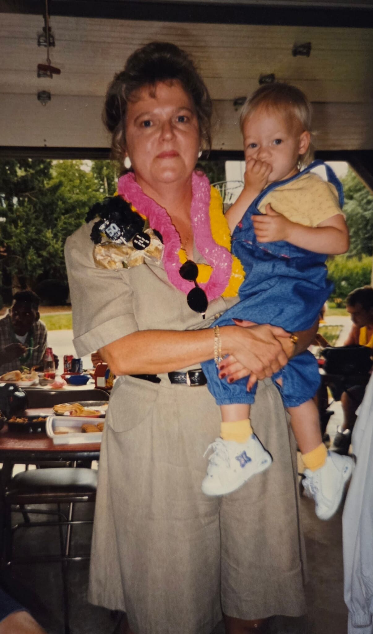 Woman in a tan dress holding a toddler at a gathering with a buffet table in the background, wearing a pink and yellow lei.
