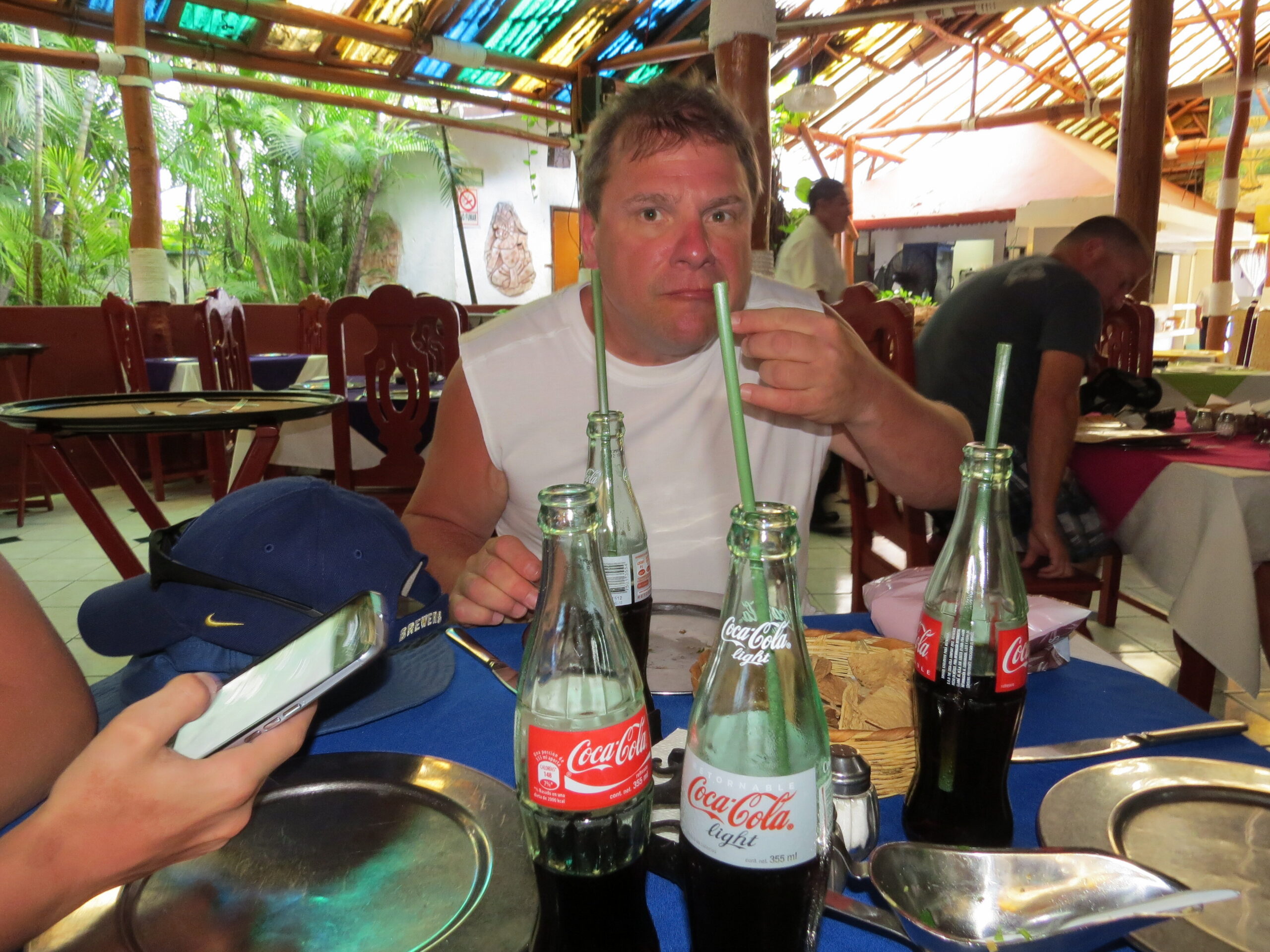 Man in a white sleeveless shirt at a table with multiple Coca-Cola bottles and green straws in an open-air tropical restaurant setting.
