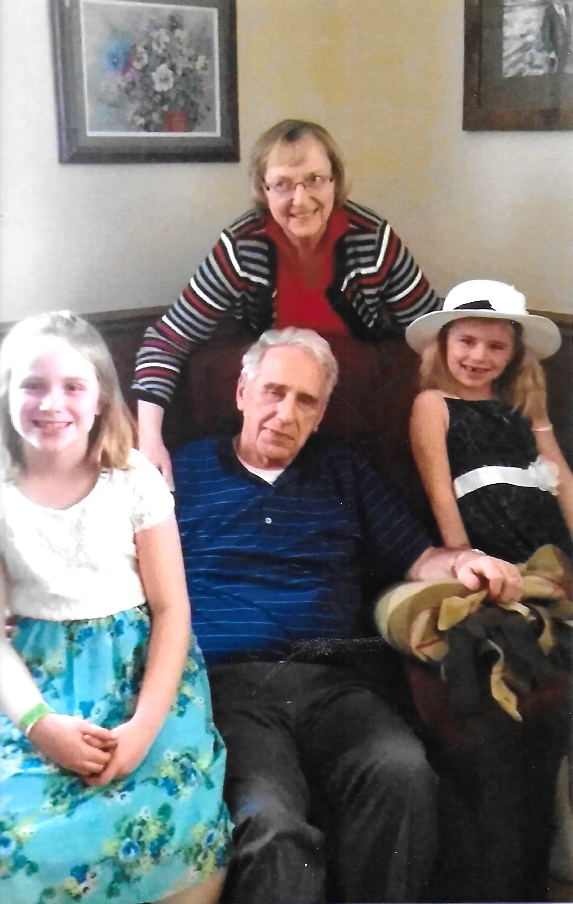 Smiling elderly woman in a striped cardigan leaning over a couch behind a seated man and two girls on either side of him on a sofa indoors.