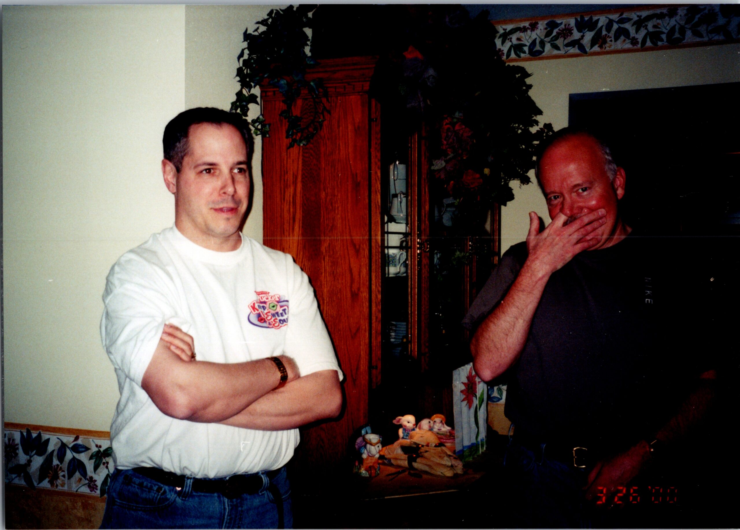 Two men stand indoors near a wooden cabinet with plants; one crosses his arms in a white T-shirt, the other covers his mouth with his hand.