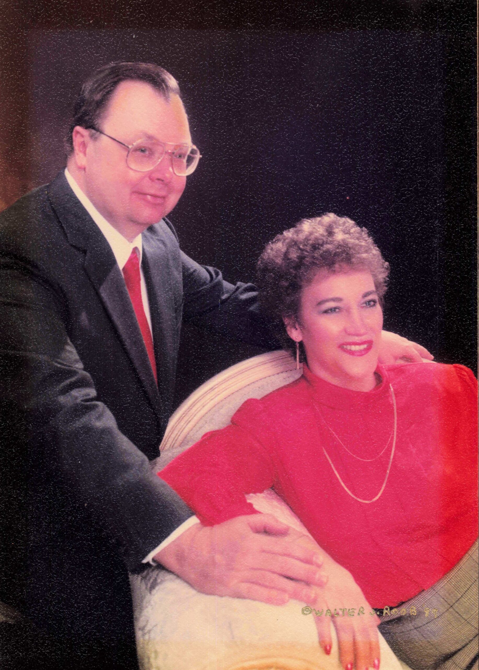 Man in a dark suit with red tie sits beside a smiling woman in red, posed on a striped couch (vintage studio portrait).
