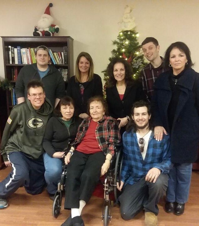 Group of ten people posing for a Christmas photo around a decorated tree; elderly woman in a wheelchair center, family gathered.