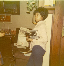 Person standing in a living room holding a newspaper and a small camera/icon in their hands, with a chair and shelves in the background.
