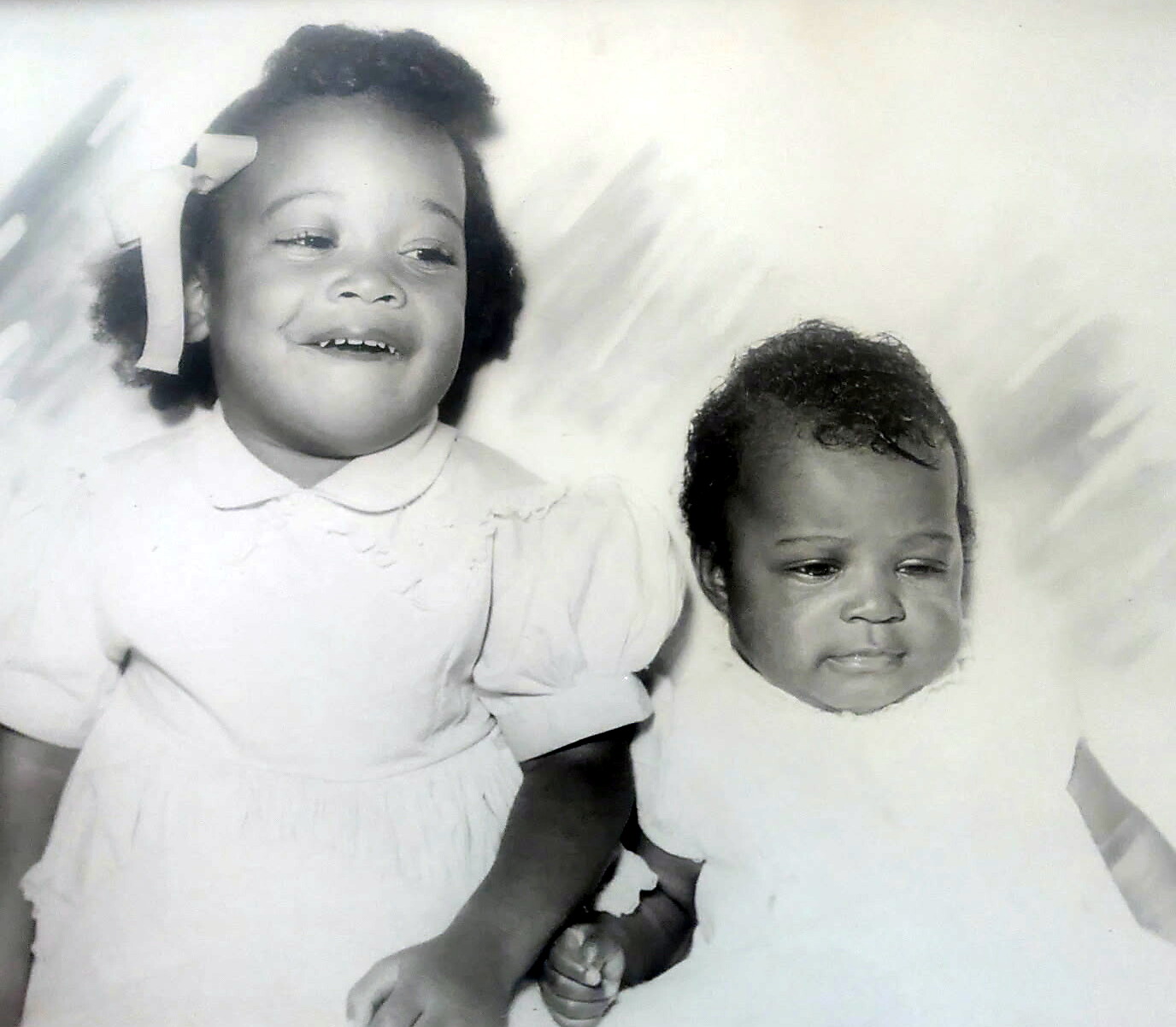 Two young siblings posing together: a smiling girl with a bow in her hair beside a baby.