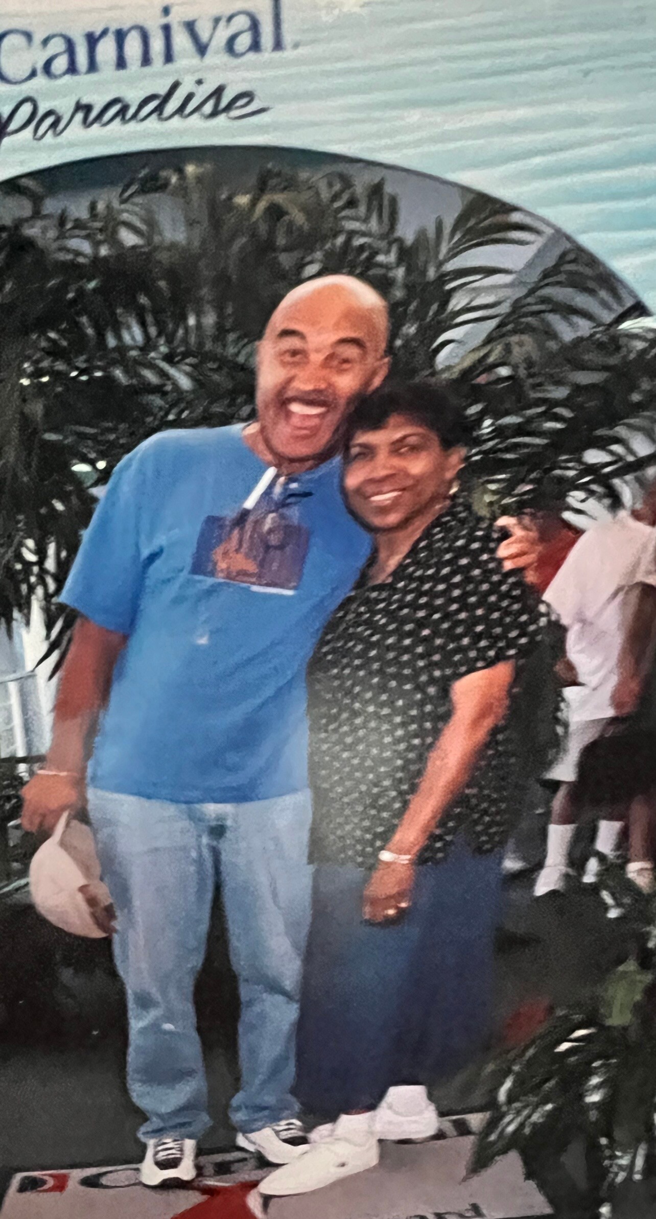 Happy couple posing together, man in blue T-shirt and jeans, woman in a black polka-dot blouse, tropical plants in the background. 'Carnival Paradise' sign is visible at top edge.