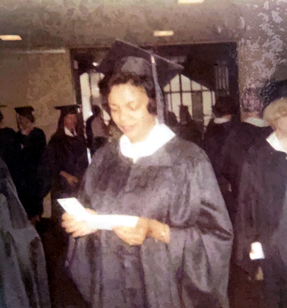 Graduate in a black gown and cap reading a booklet at a commencement ceremony, with other graduates in robes in the background.