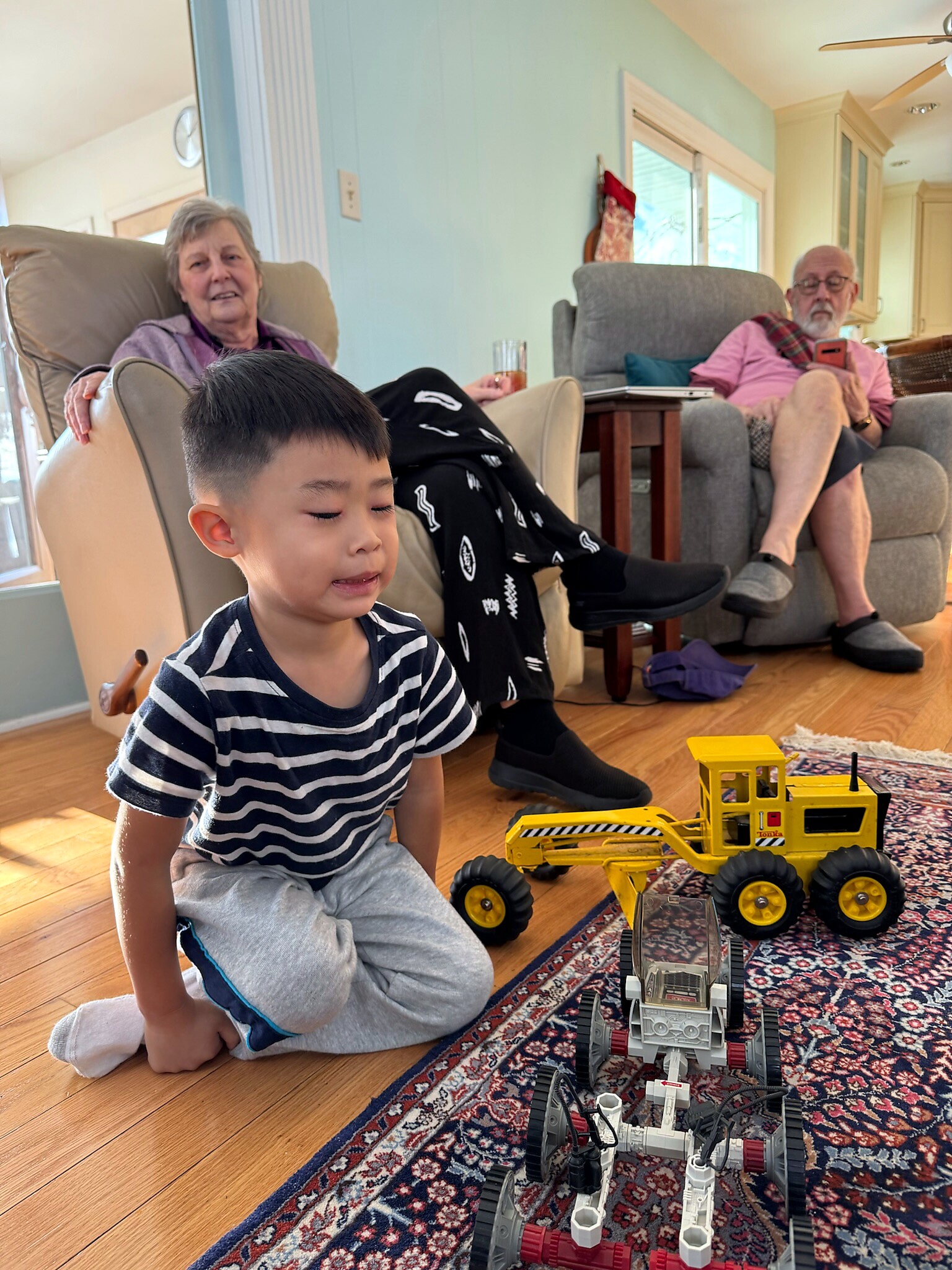 A young boy sits on the floor playing with yellow toy construction trucks while two seniors relax in chairs in a bright living room.