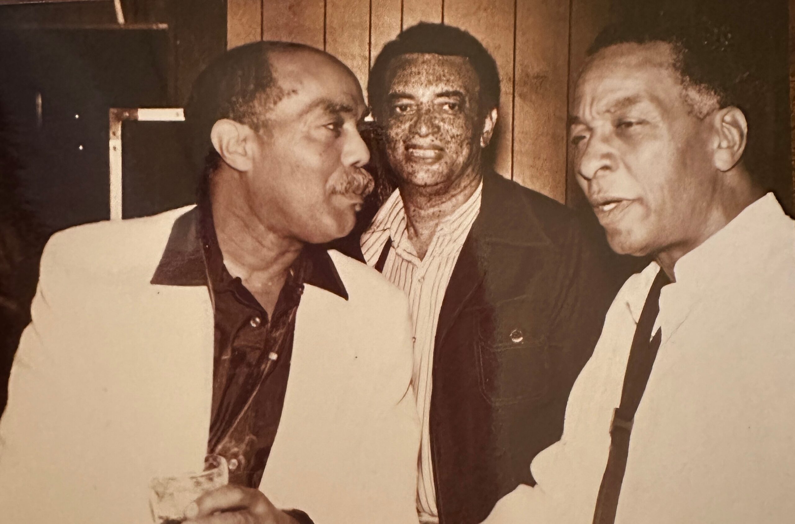 Three men in vintage outfits posing closely for a group photo against a wood-paneled background.