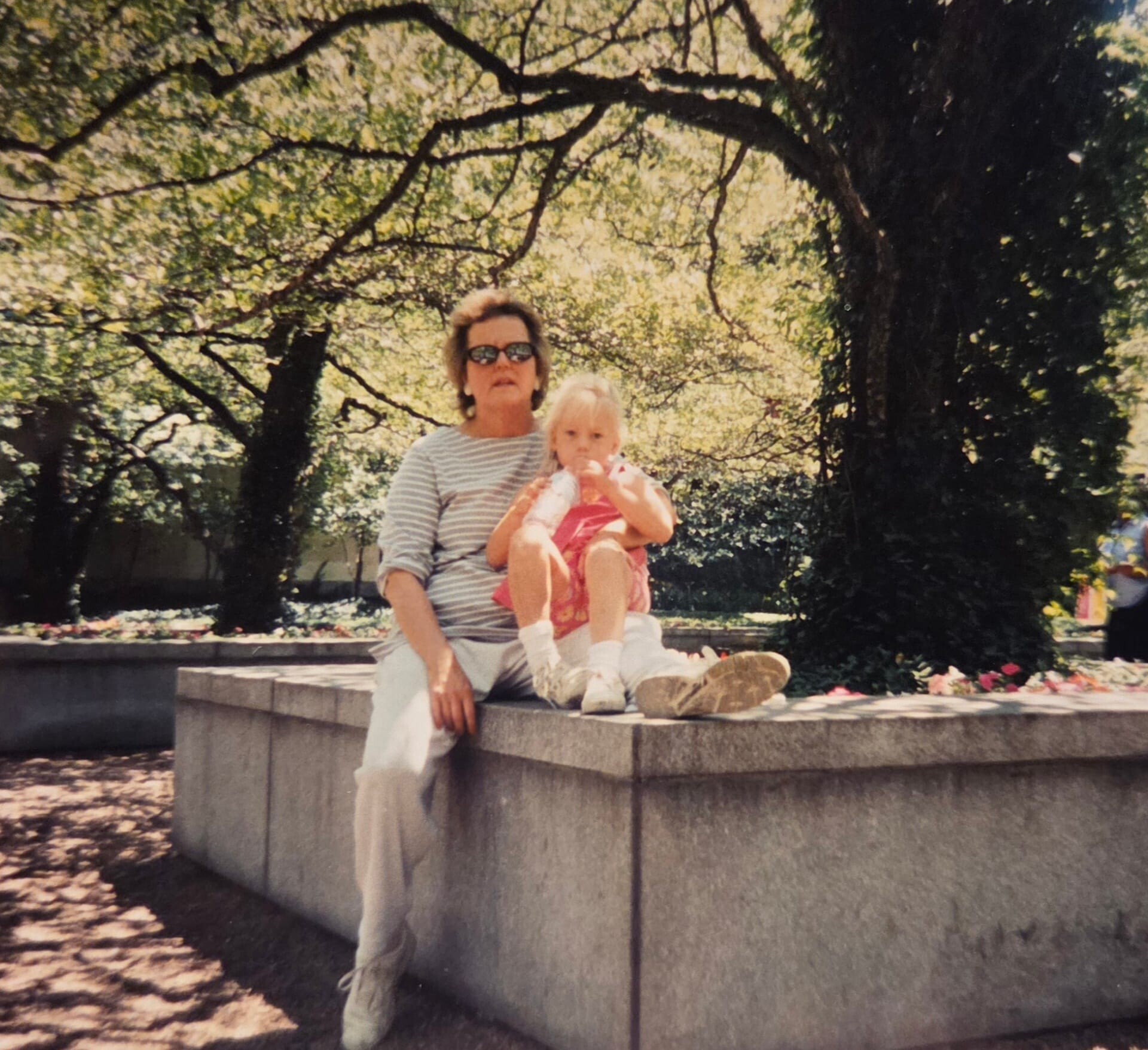 Woman in a striped shirt and sunglasses sits on a concrete ledge holding a young child in a park with trees in the background.