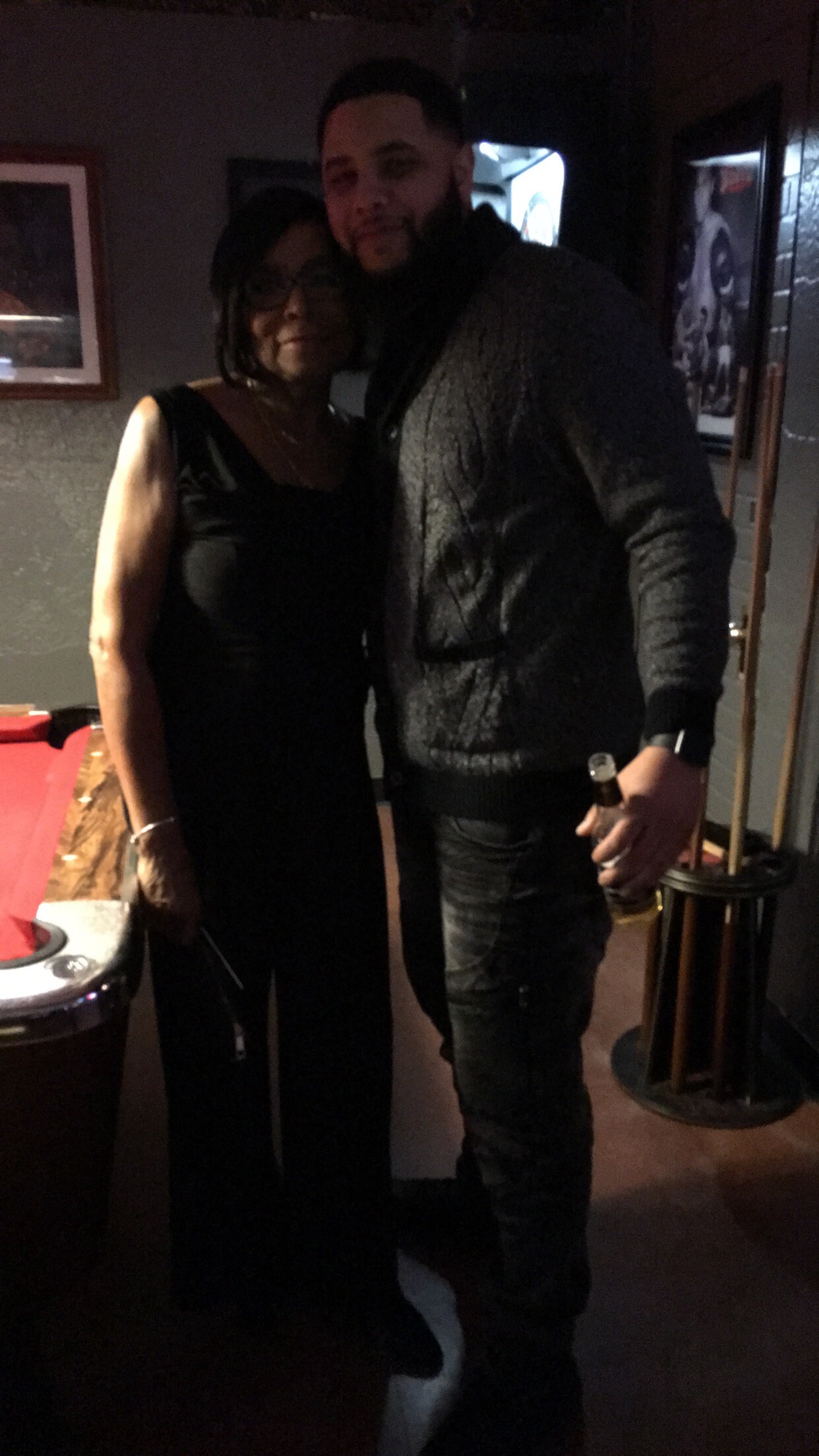 Two people posing together in a dim room; a woman in a black dress stands beside a man in a dark sweater, the man holding a beer bottle.