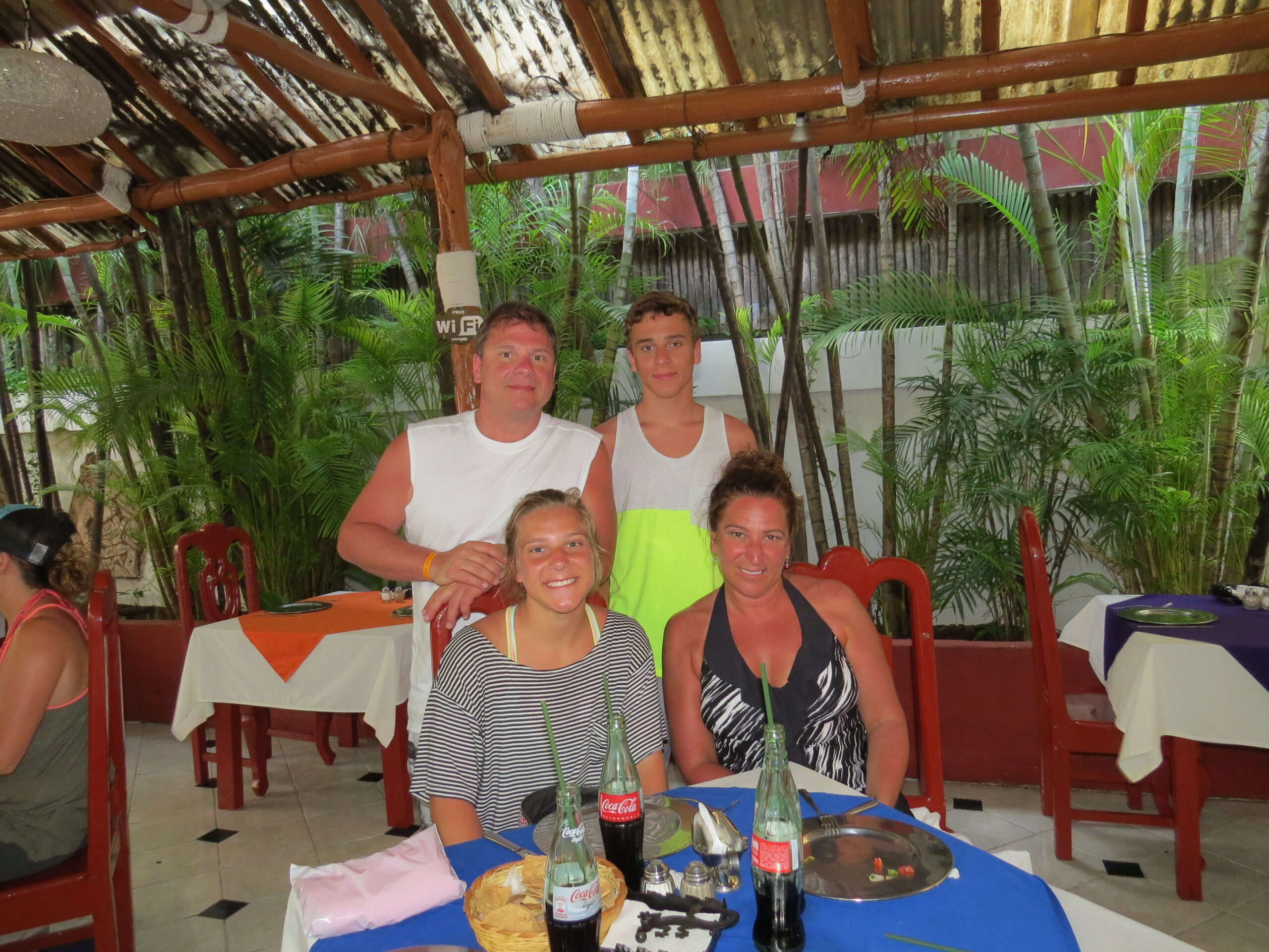 Group of four people posing at an outdoor tropical restaurant, with palm plants, red chairs, and drinks on the table.