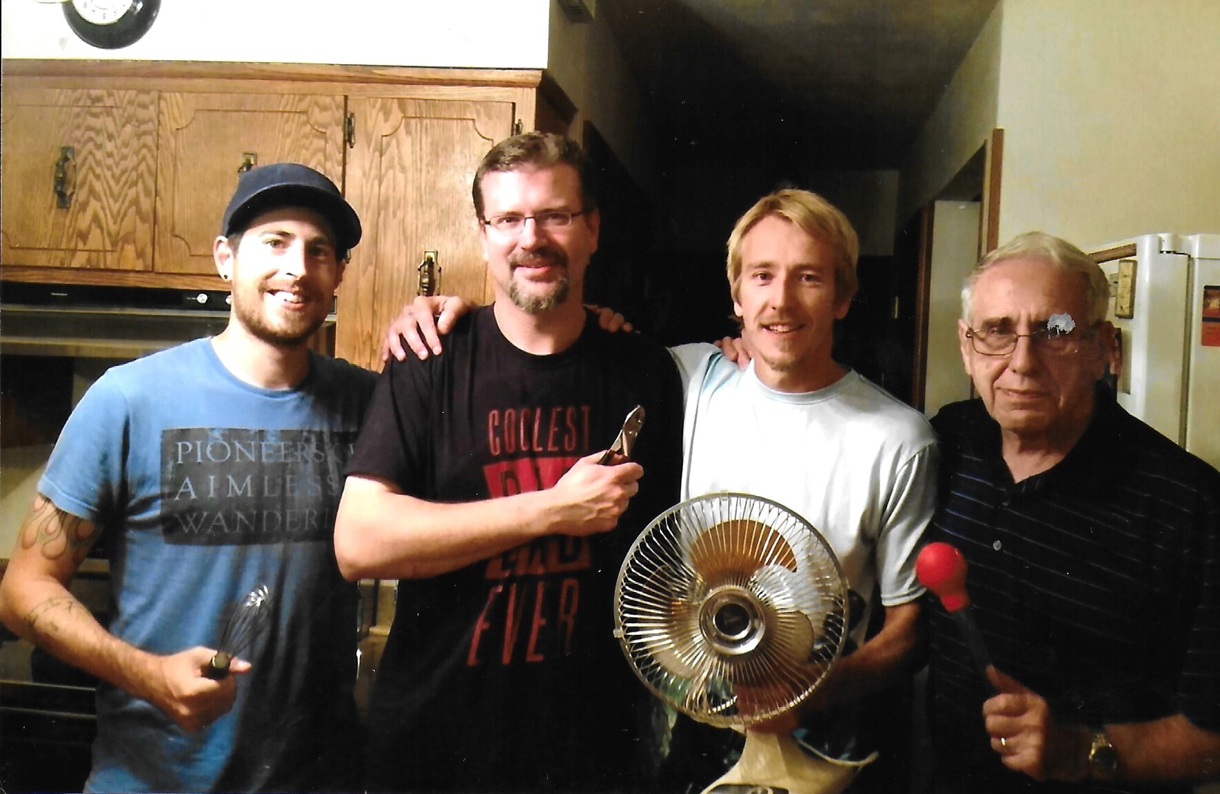 Four men posing in a kitchen, smiling, with tools and a fan in hand, suggesting a casual gathering or project time.