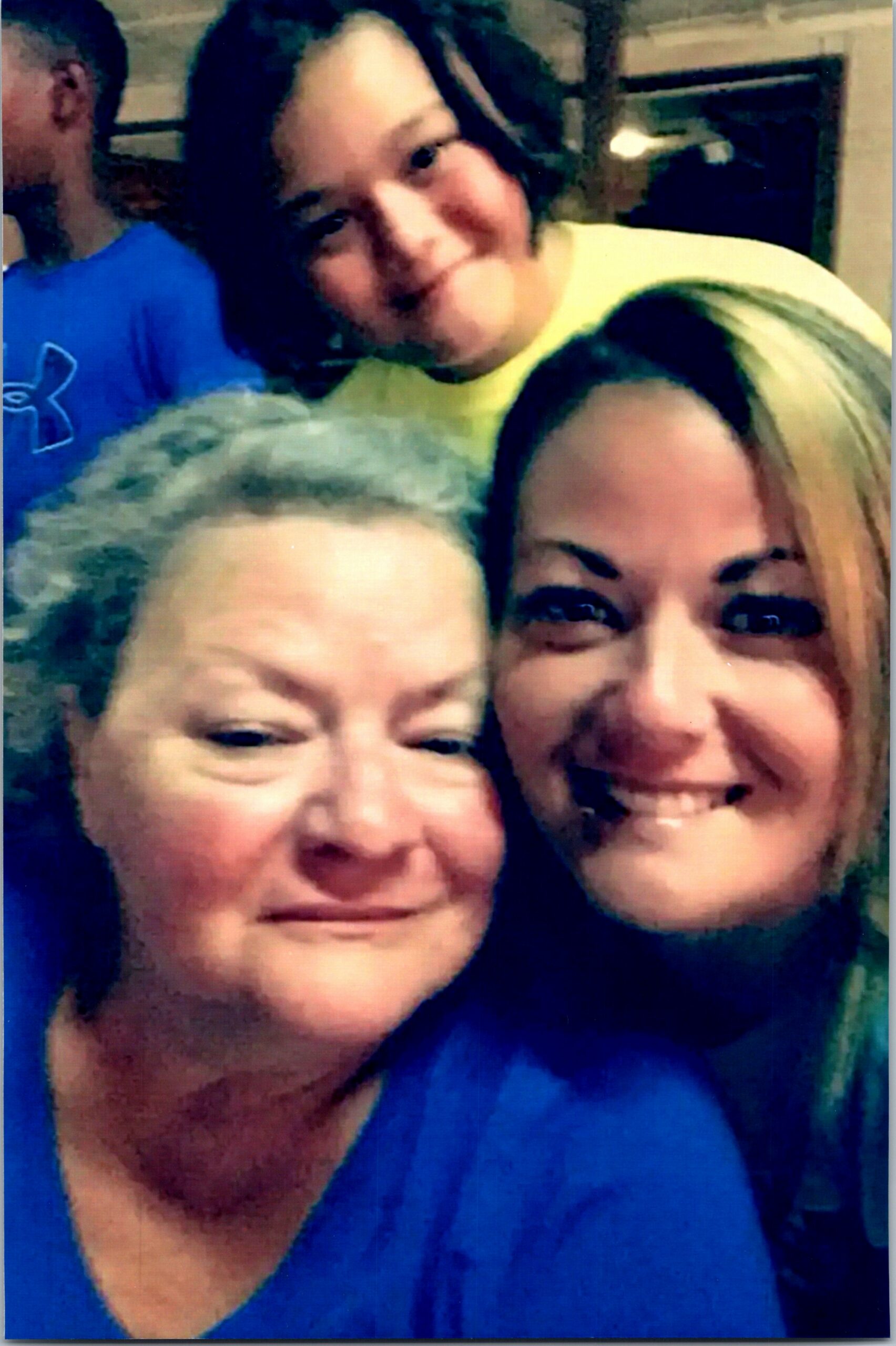 Three people smiling for a close selfie, two women in the foreground wearing blue shirts and a friend in a yellow shirt behind them, indoors.