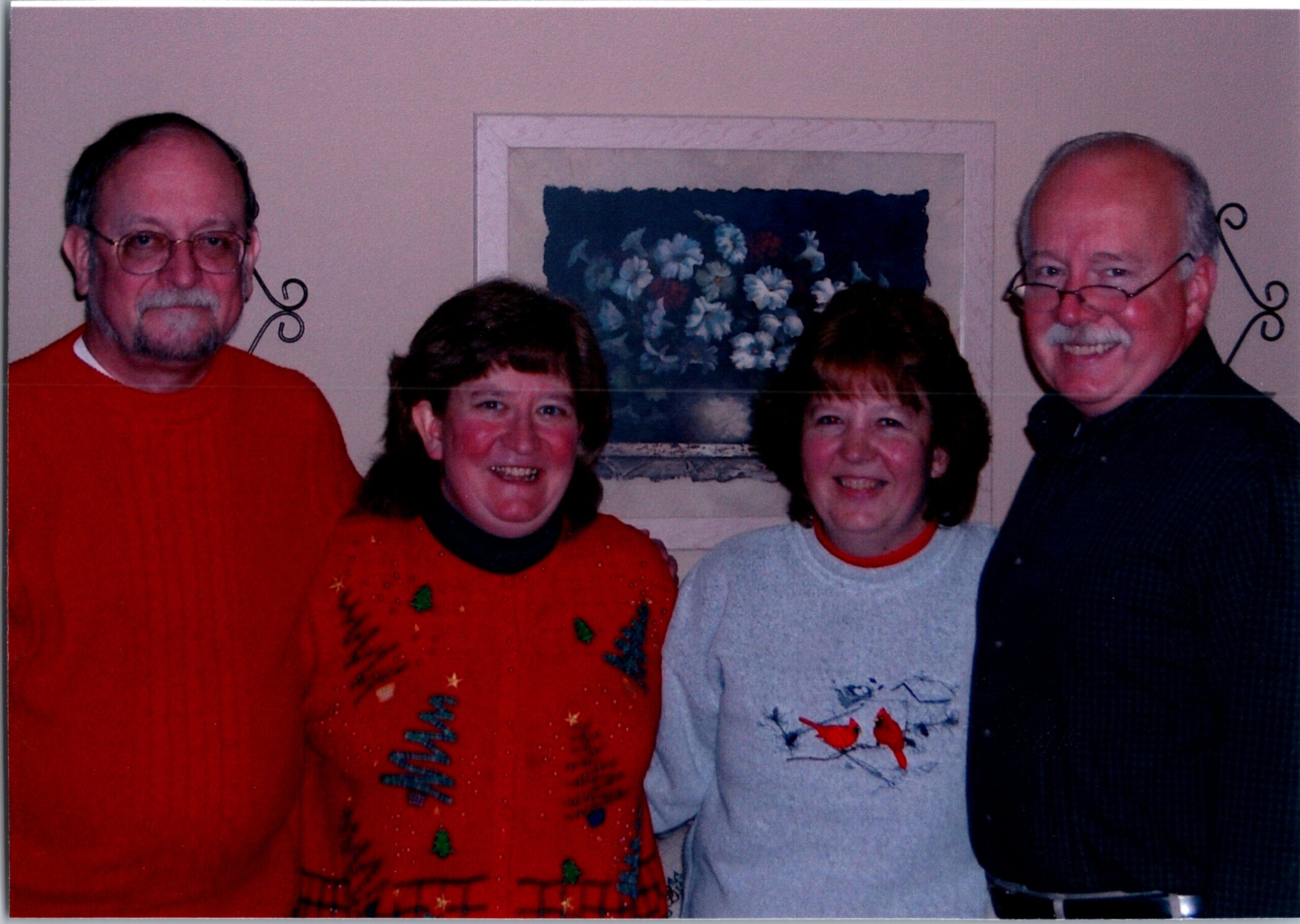 Four adults posing together indoors for a group photo; two wear red holiday sweaters and the others wear festive tops, standing in front of wall art.