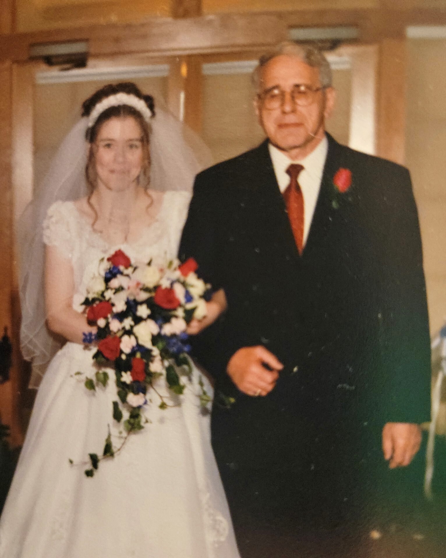 Bride in white wedding dress holding a colorful bouquet, standing beside a suited man in a dark suit and red tie at a doorway indoors.