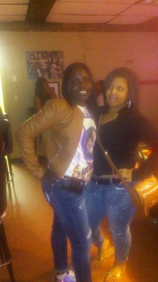 Two smiling women pose together in a dimly lit room, wearing jeans; one in a brown jacket and graphic tee, the other in a black top with a crossbody bag.