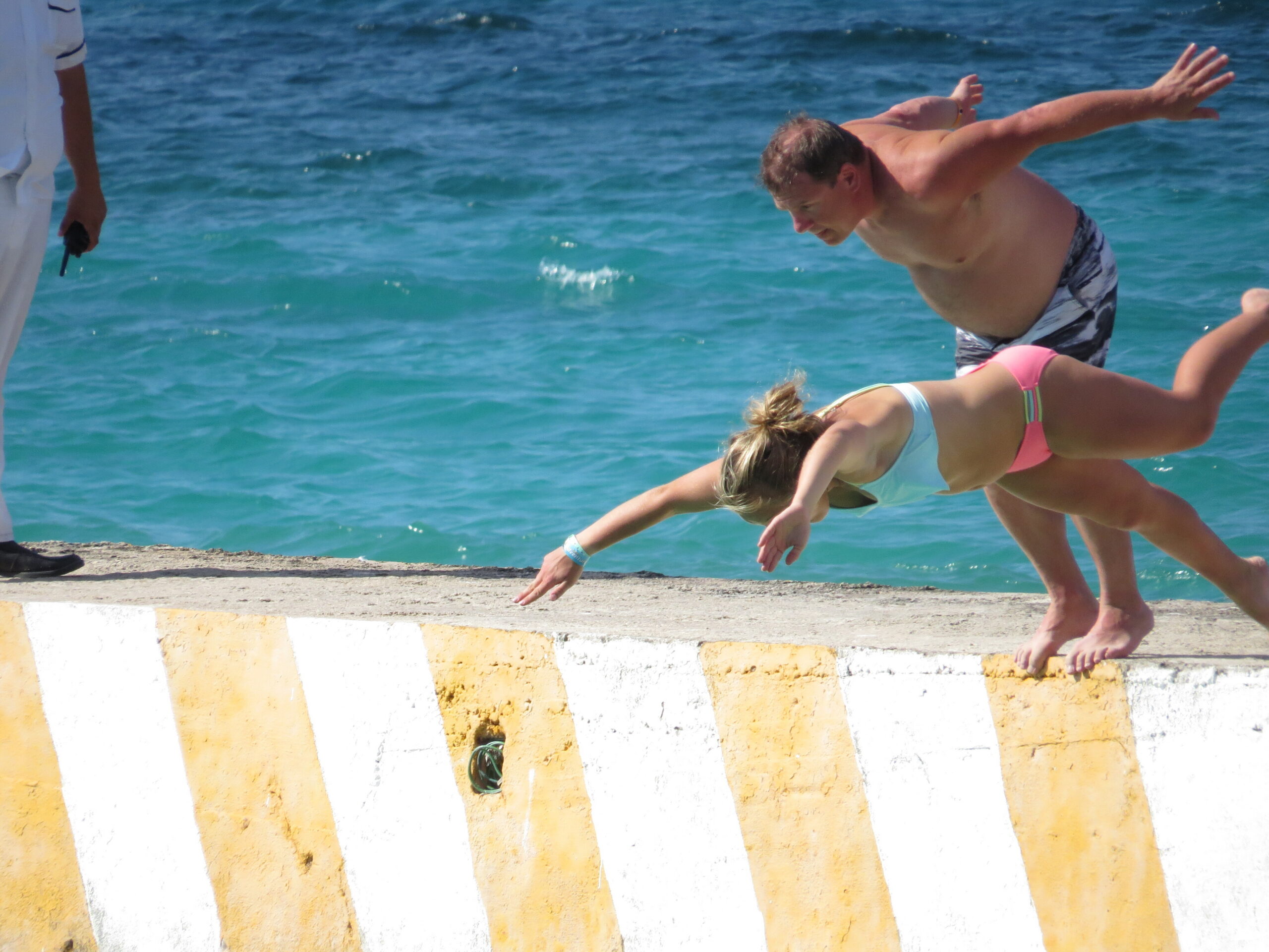 Two swimmers leap off a yellow-and-white striped seawall into the turquoise sea, arm extended, in bright beachwear; a person in white stands to the left holding something.