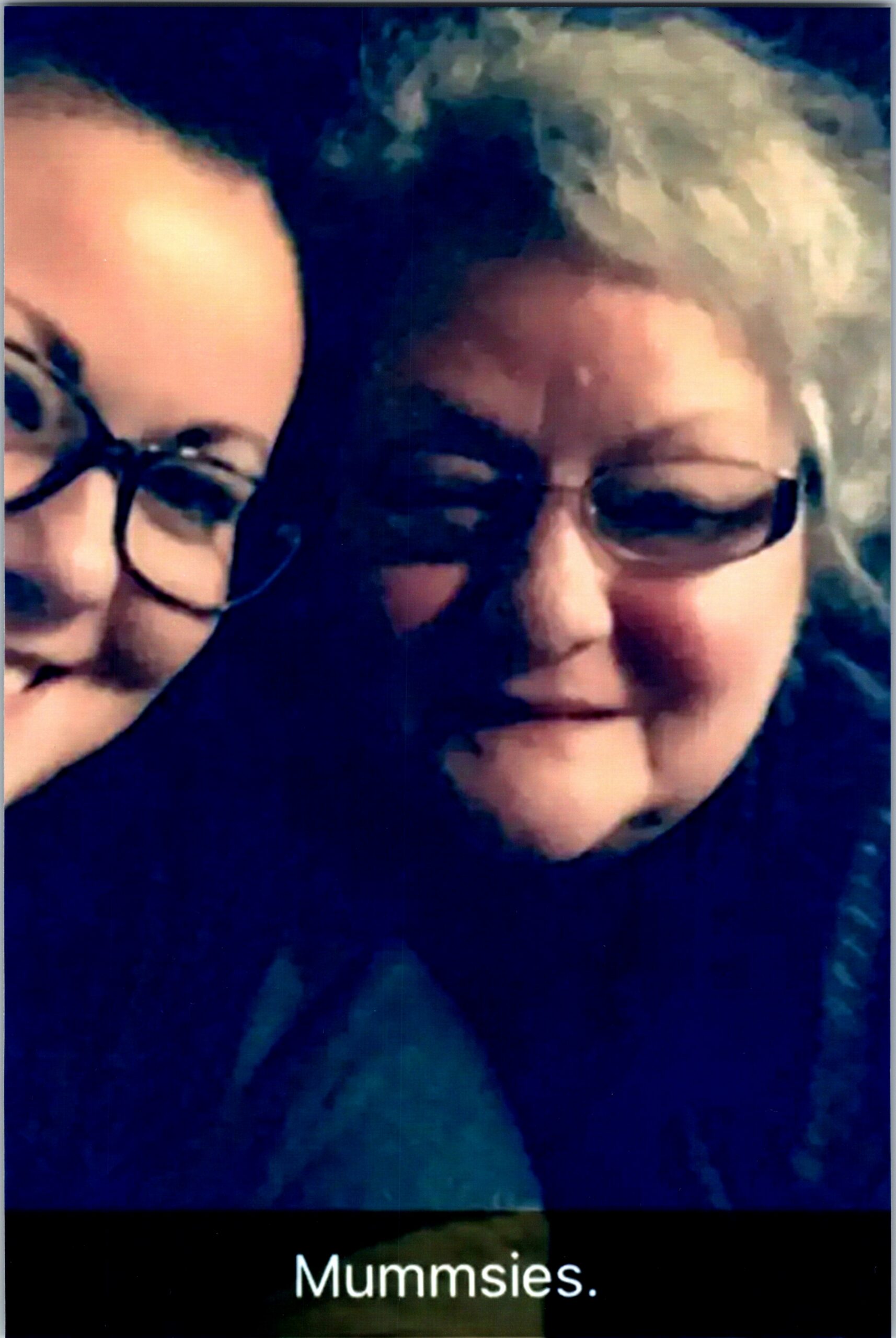 Close-up selfie of two smiling people wearing glasses, cropped faces in a dark blue background; bottom shows the word 'Mummisies'.