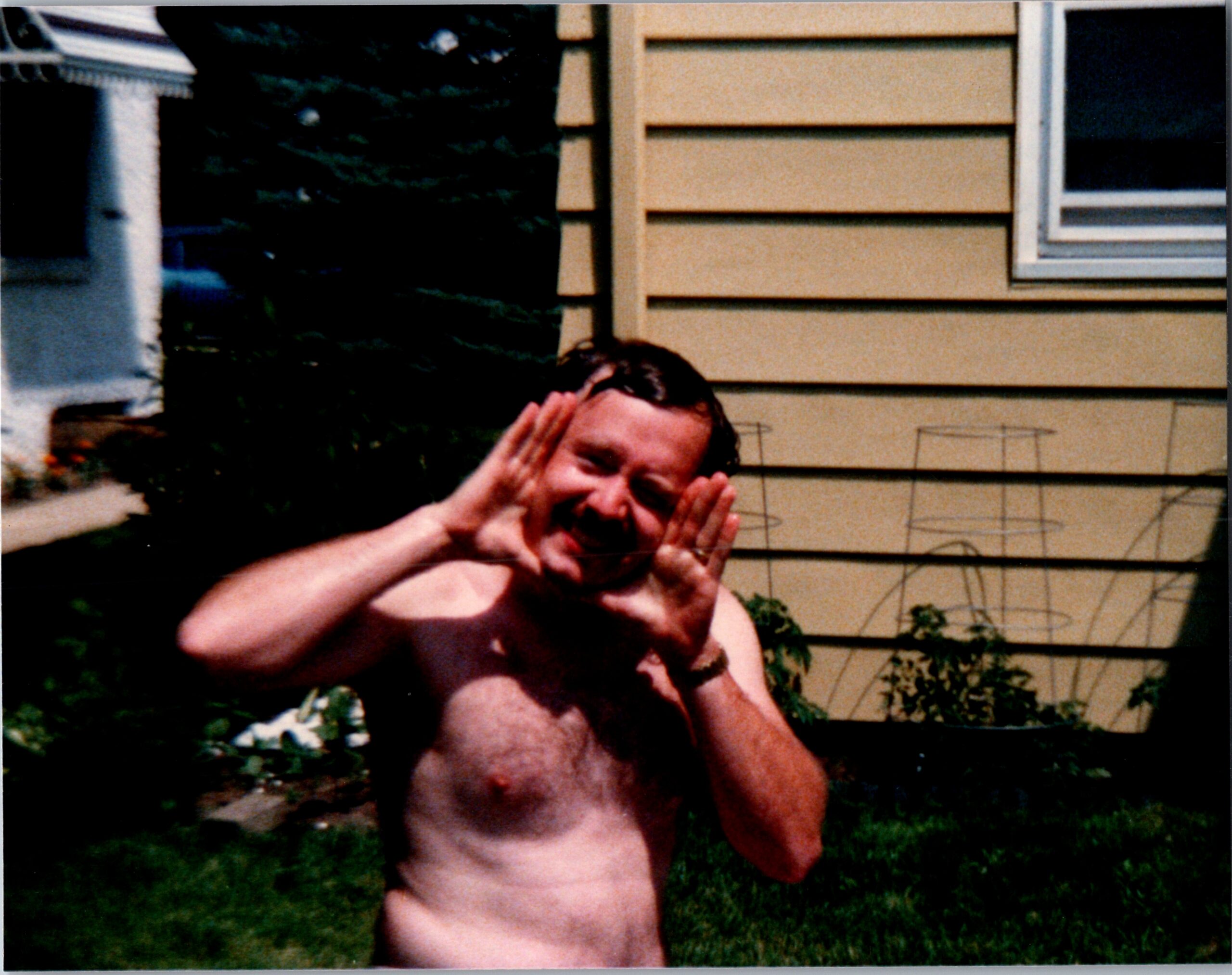 Shirtless man outdoors, hands near his face in a playful pose, with a beige house and green lawn in the background.