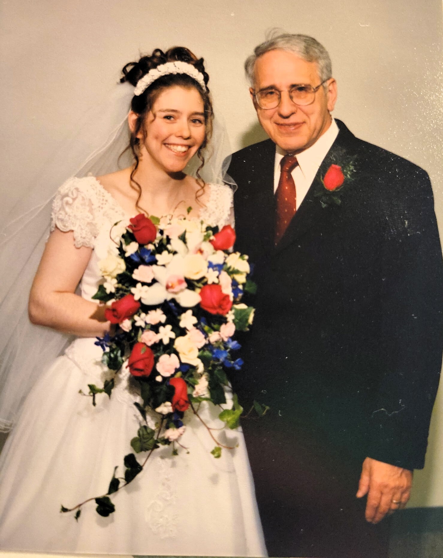 Bride in a white lace wedding dress holding a colorful bouquet, smiling beside an older man in a dark suit with a red boutonniere and tie.