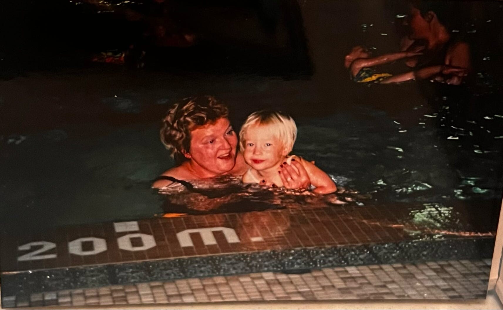 Woman and young child swimming together in a dark pool, smiling, near the pool edge with a '200 m' marking visible on the tiles.