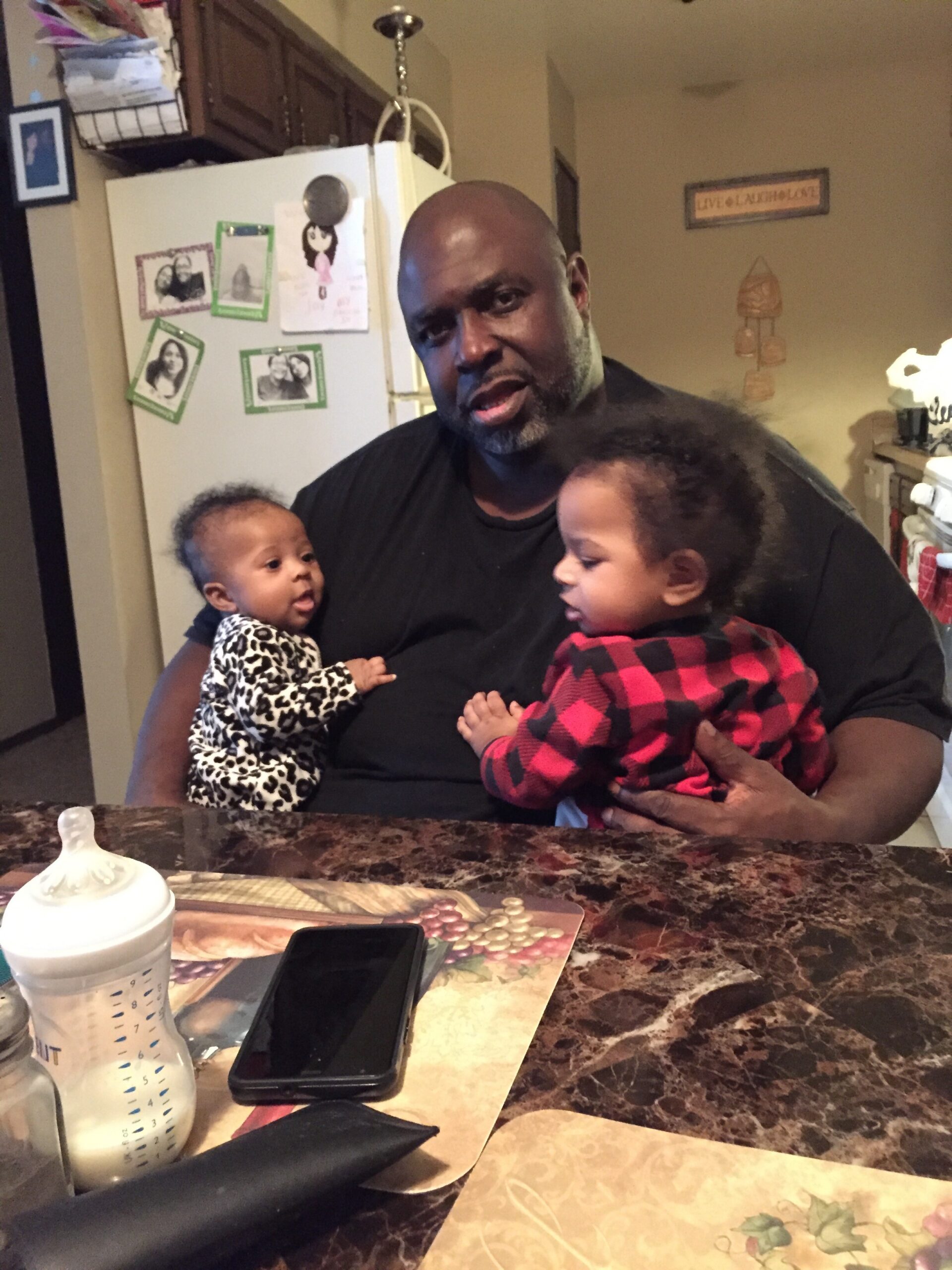 A man in a black shirt holds two babies, one in each arm, in a kitchen.