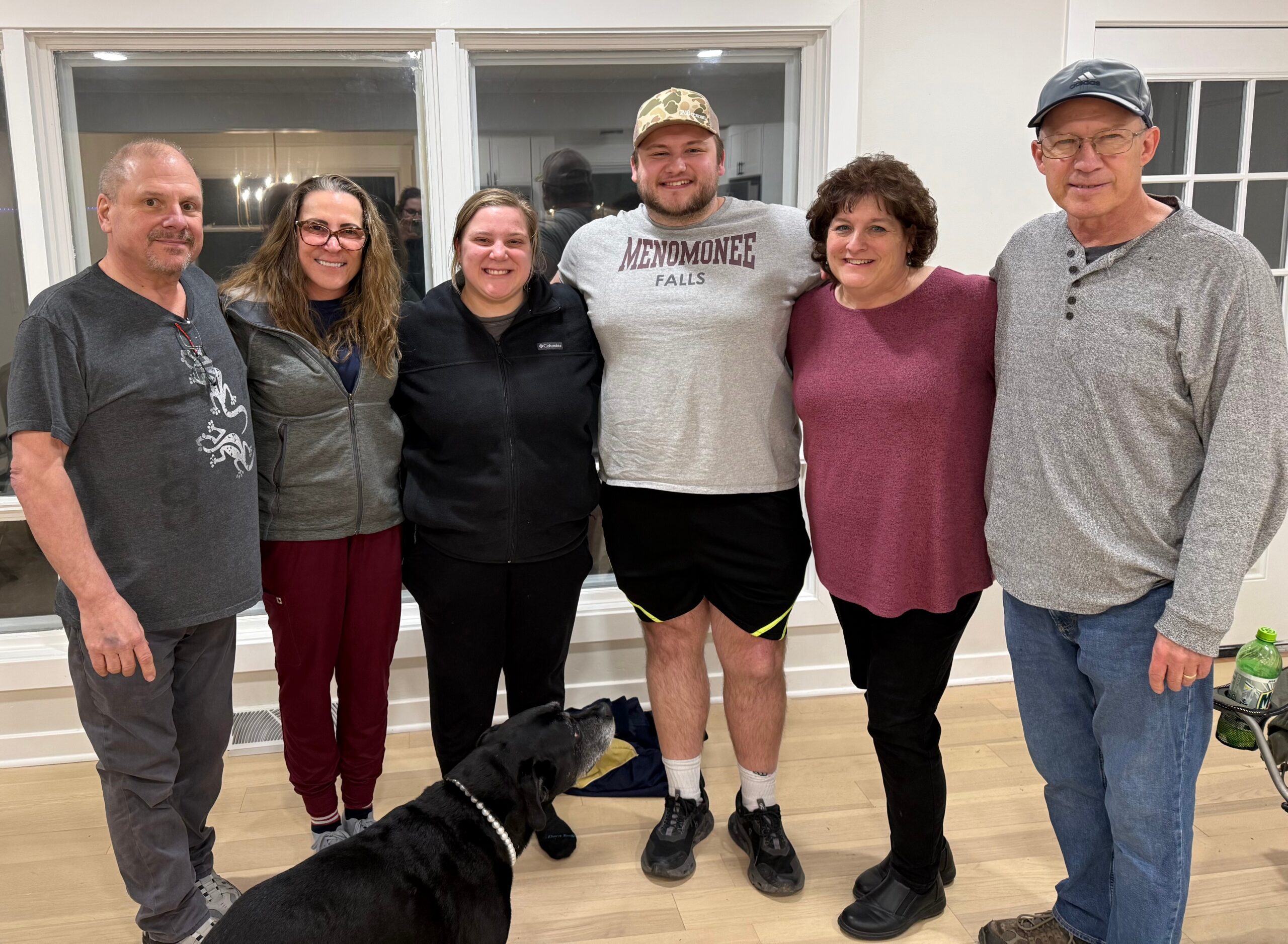 Group of six adults posing indoors, smiling at the camera with a black dog at their feet in front of them.