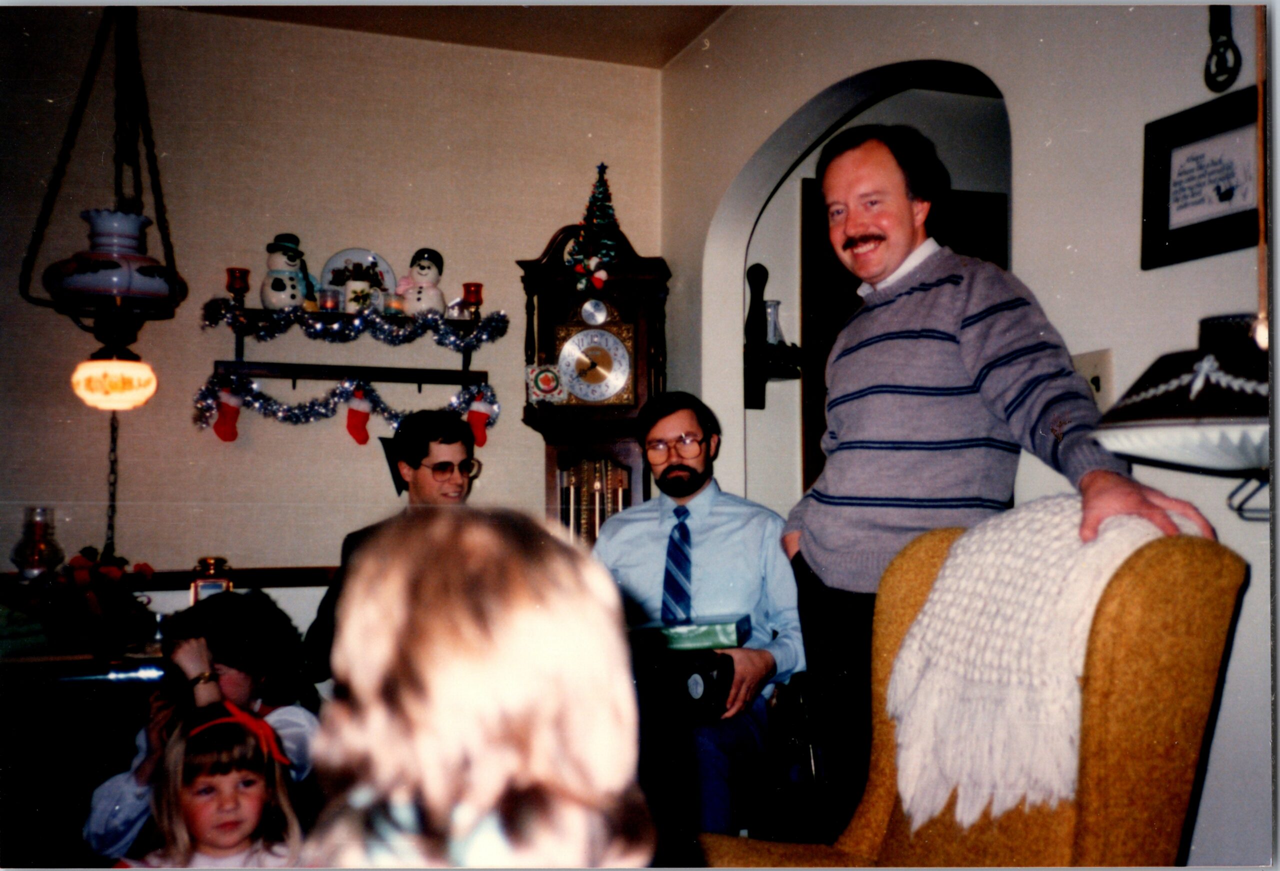 Indoor Christmas party with a man in a striped sweater standing and smiling, others seated, decorations include garlands, snowmen, and a clock.