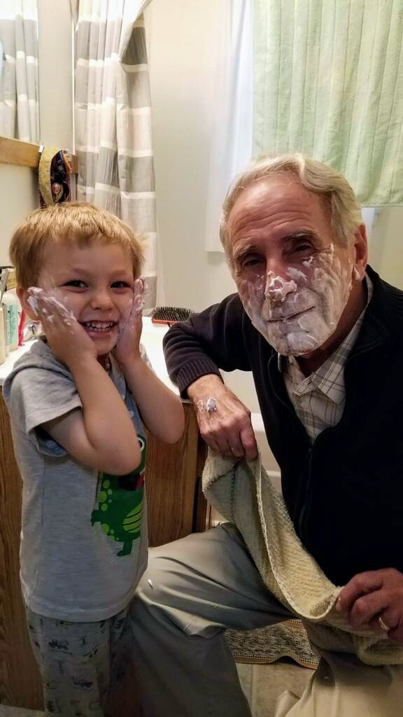 Young boy and an older man with shaving cream on their faces, smiling in a bathroom with towels nearby.