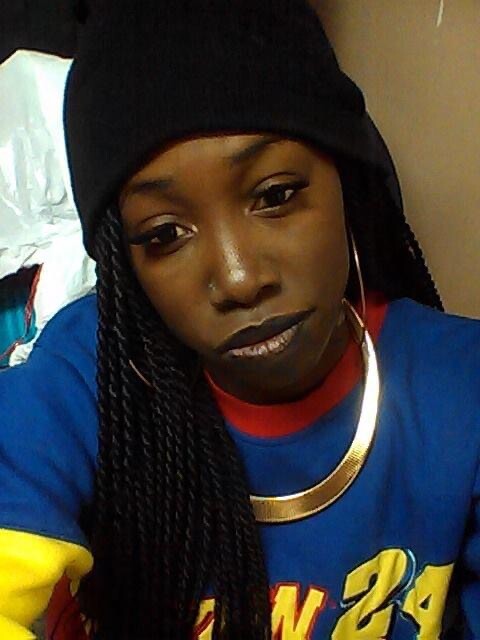 Portrait of a woman with long braids wearing a black beanie, a blue shirt with yellow accents, and a gold necklace, looking at the camera.