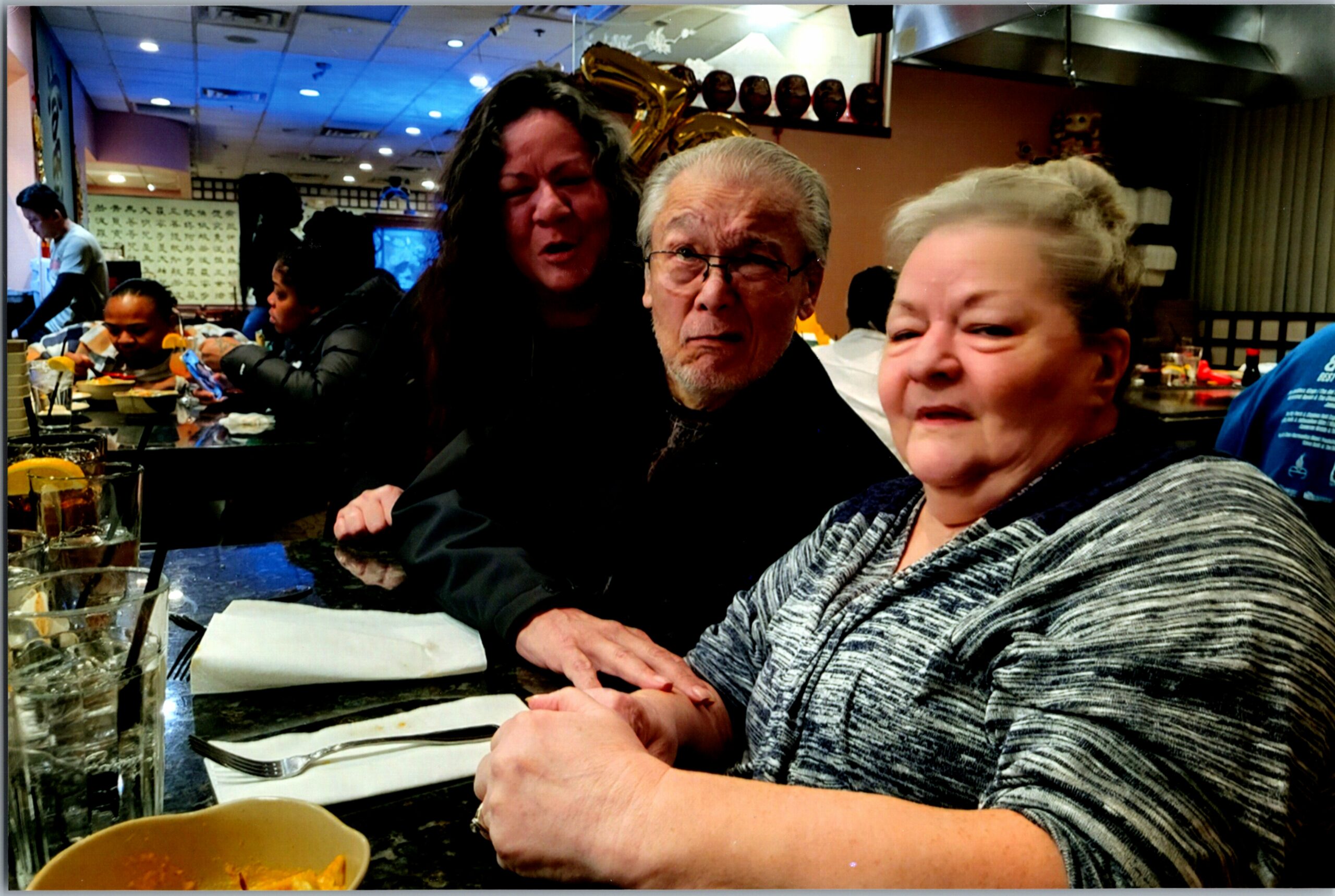 Three people sit at a restaurant table, with an older woman smiling on the right and a man with glasses center, as others in the background share a meal.