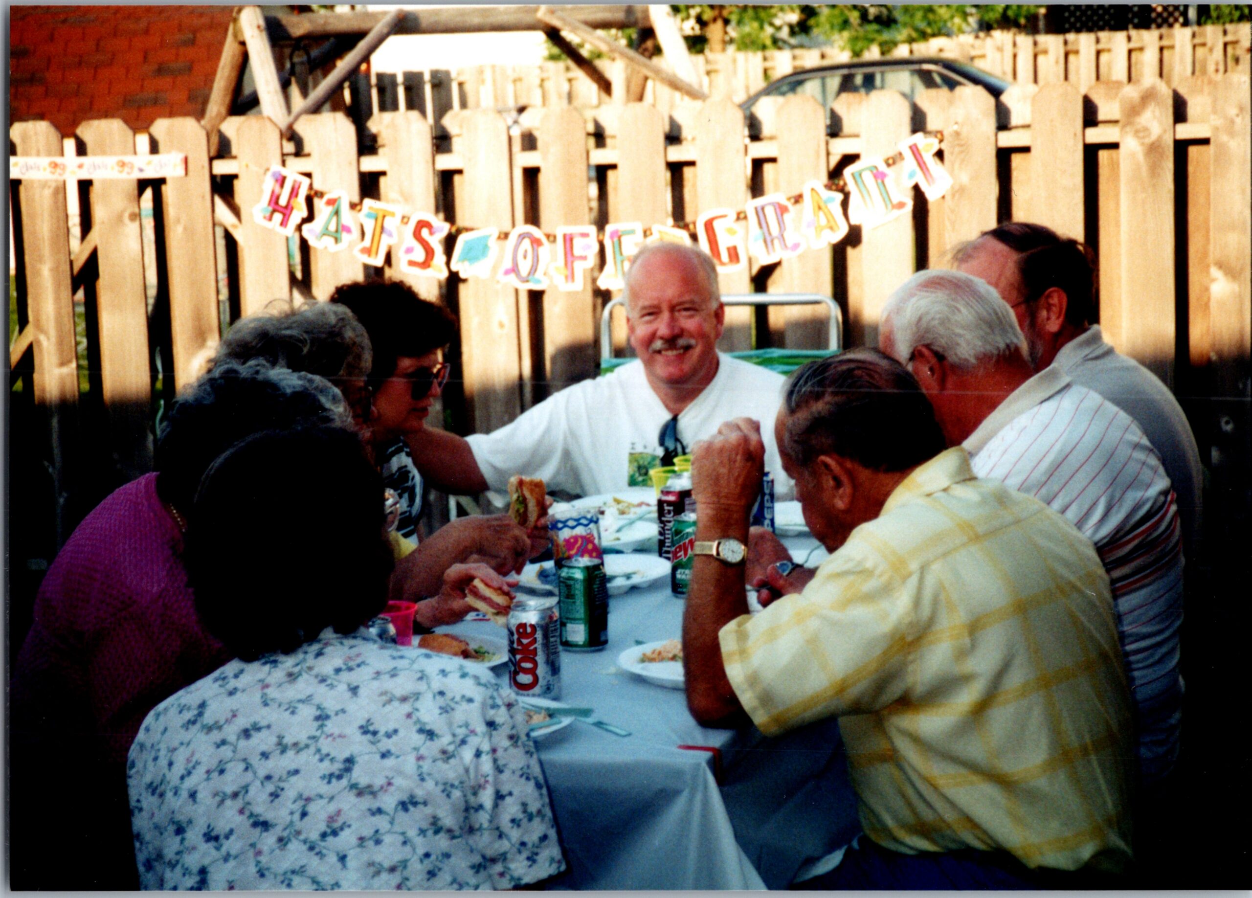 People gathered around a picnic table in a backyard, eating and drinking, with a colorful banner hanging on a wooden fence in the background.