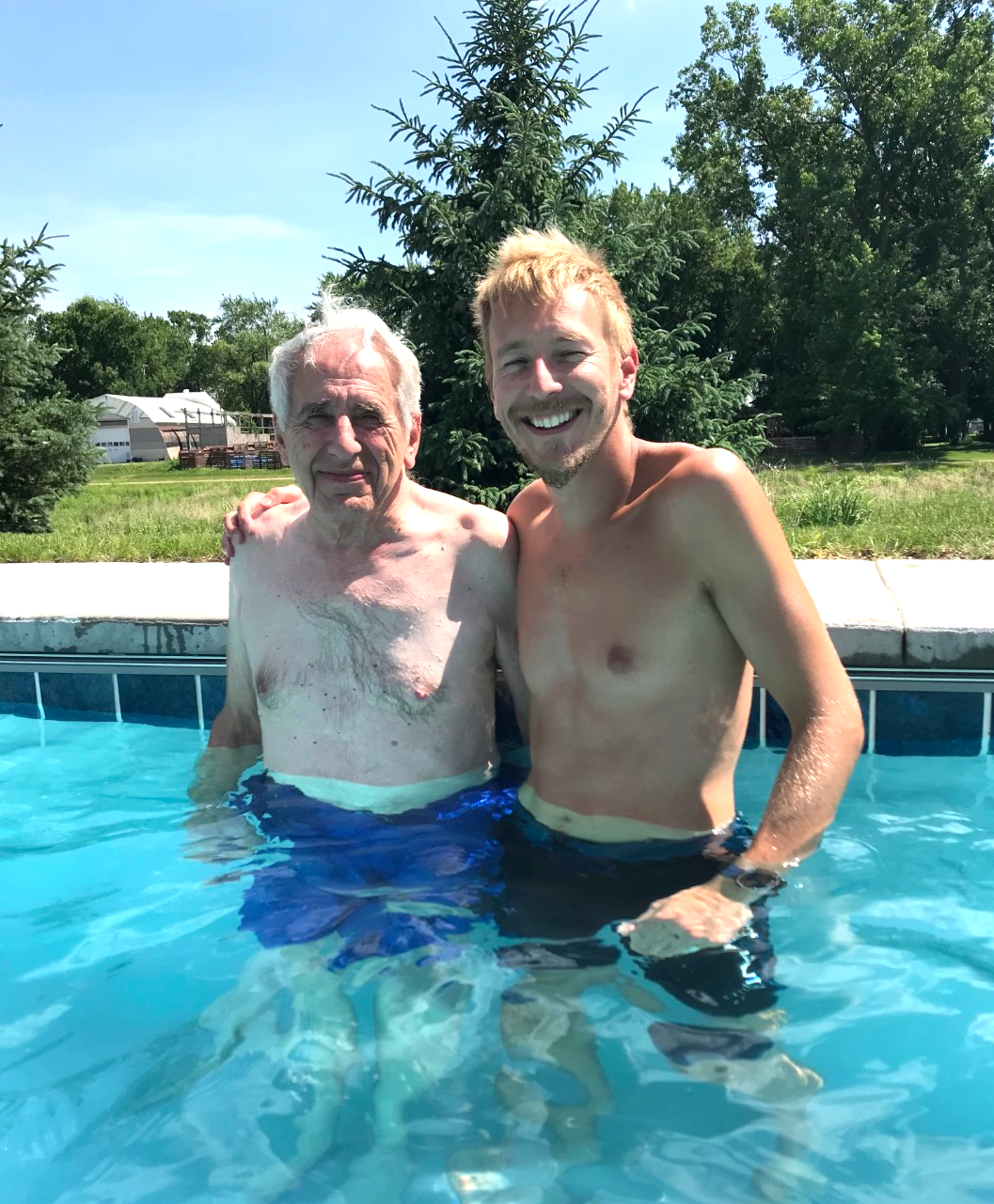 Two men stand in a backyard pool, smiling with their arms around each other; an evergreen tree and green yard behind them.