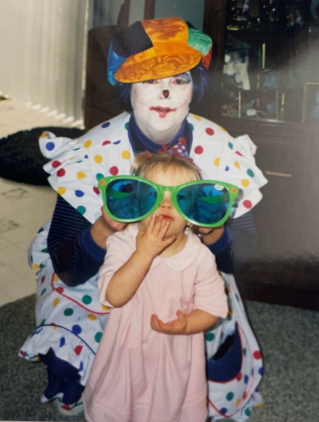 Clown in white makeup and a colorful hat poses behind a toddler wearing oversized green sunglasses.
