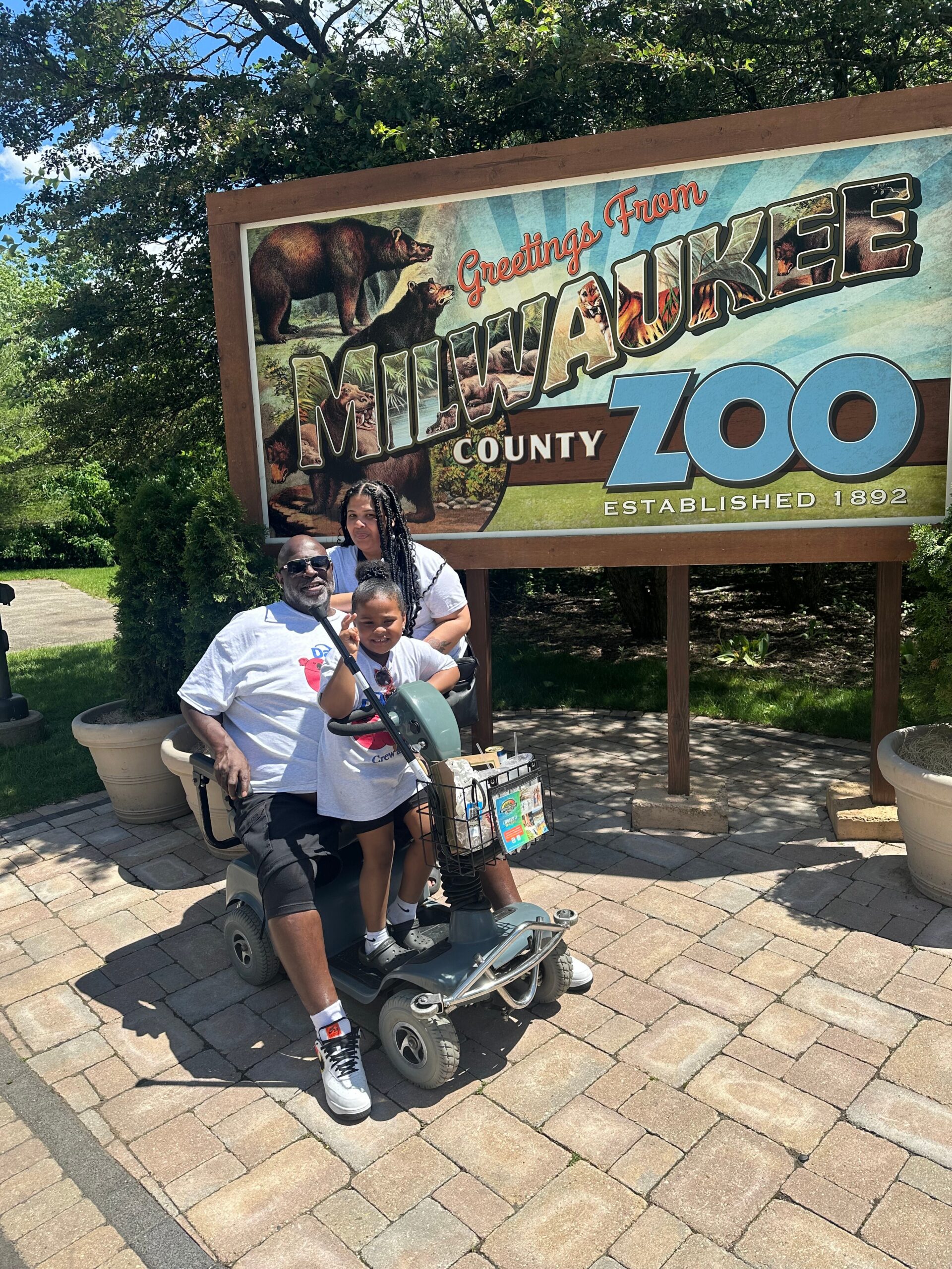 Family posing on a mobility scooter in front of a Milwaukee County Zoo sign that says 'Greetings from Milwaukee Zoo' with animal illustrations in the background.