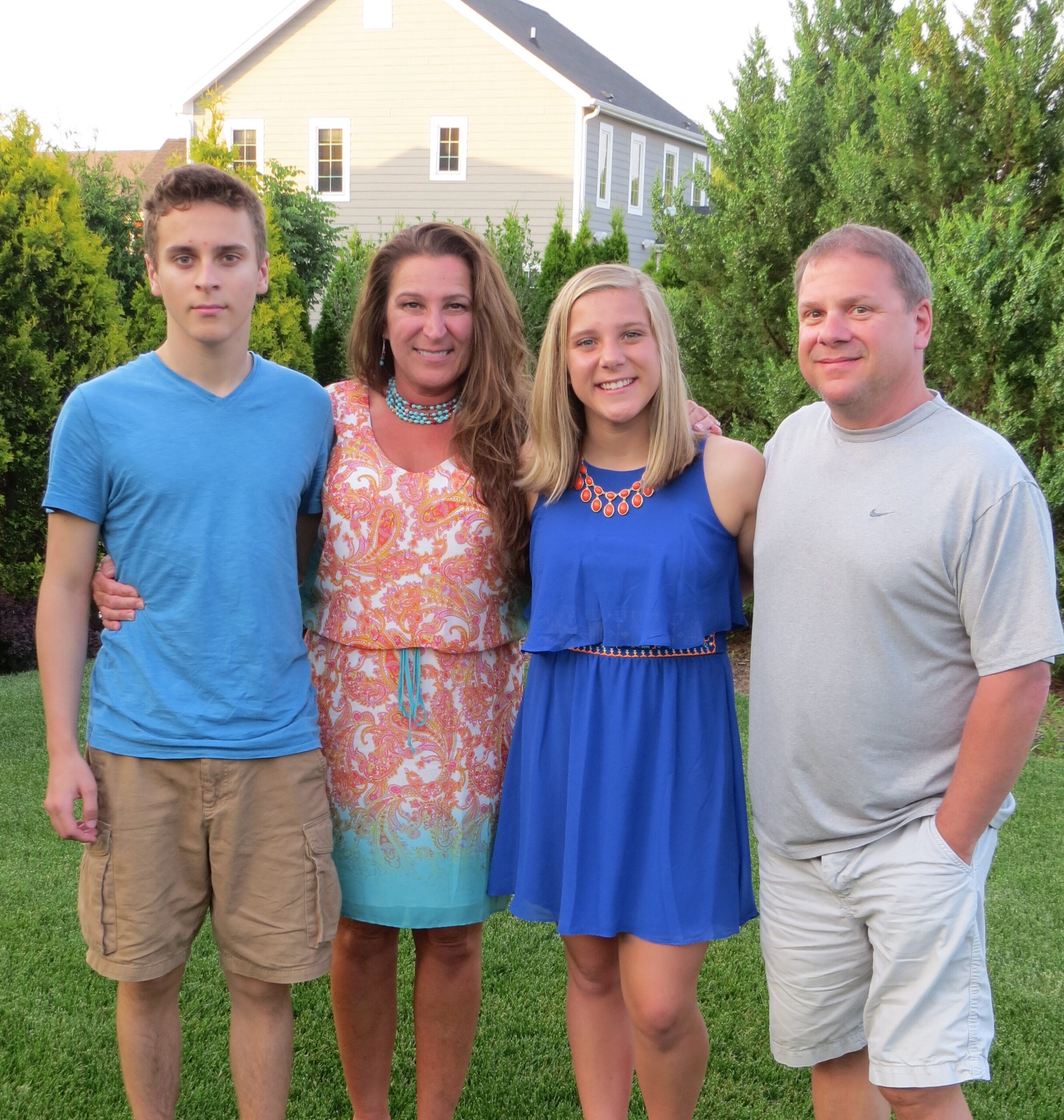 Four people stand outdoors on a green lawn with a house and trees behind them, posing for a photo.