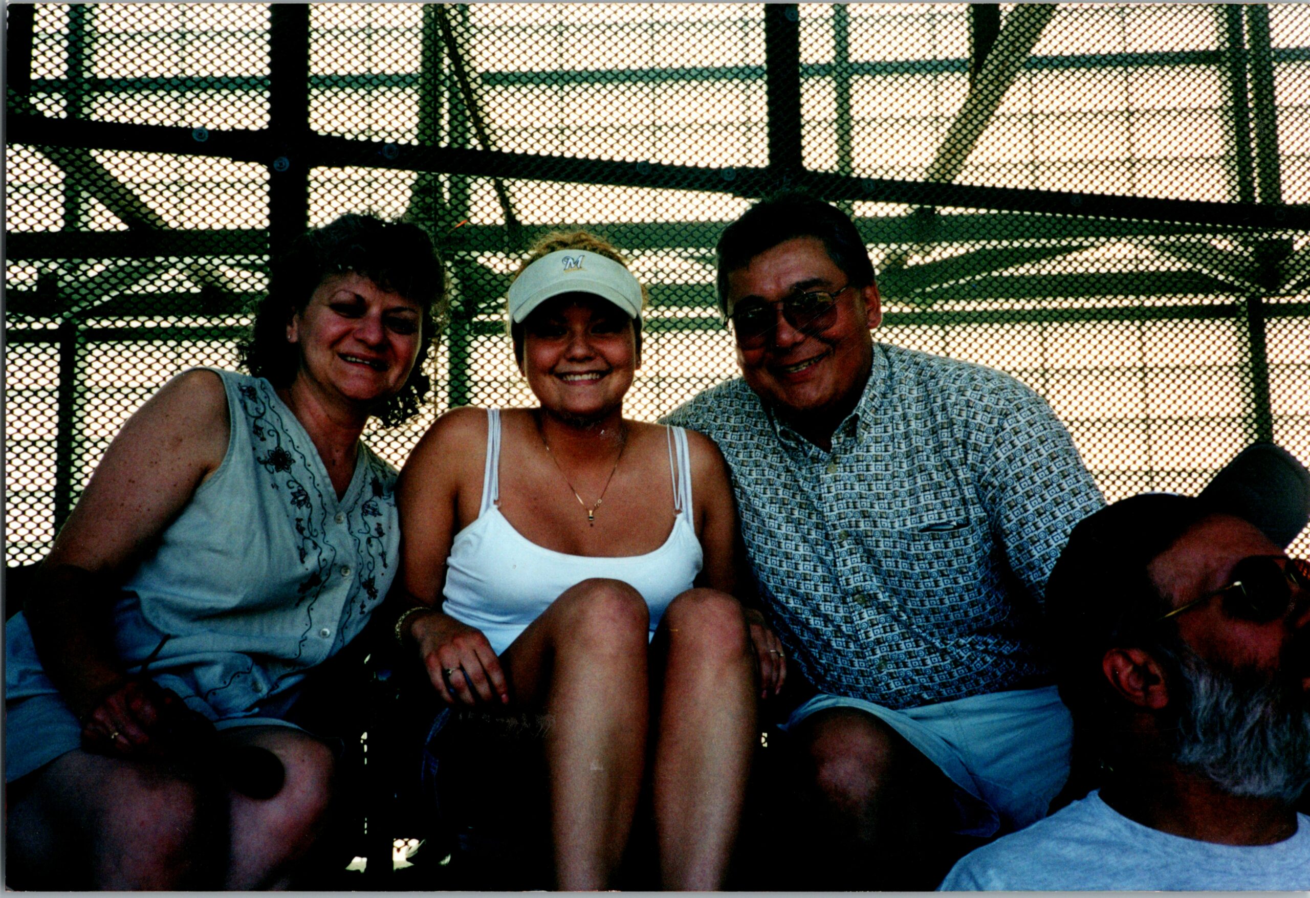 Three adults smiling and posing closely for a casual group photo behind a metal chain-link fence behind them.