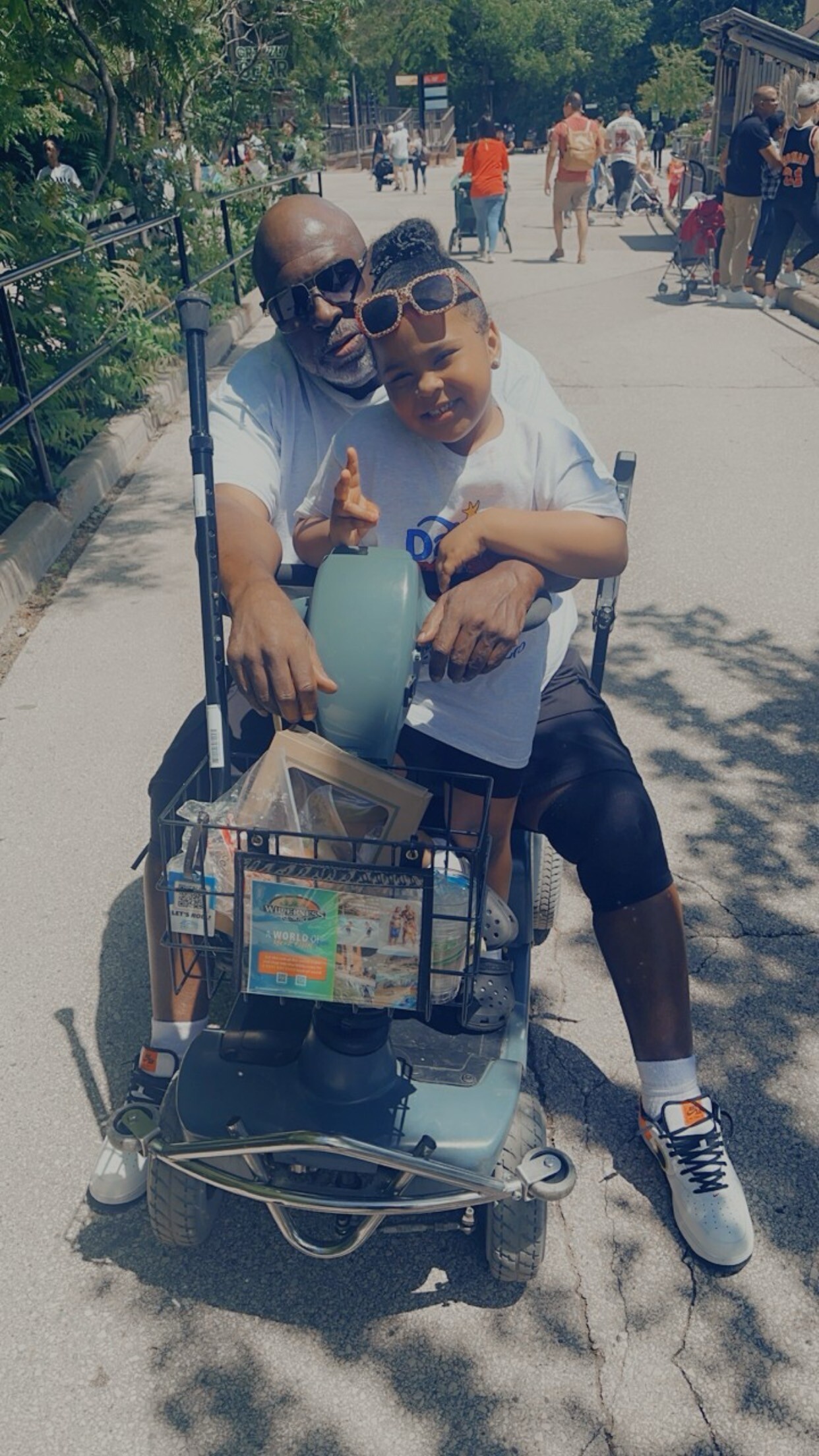 Elderly man and young girl ride a mobility scooter together on a sunny city street, smiling at the camera with a basket of items at the front.