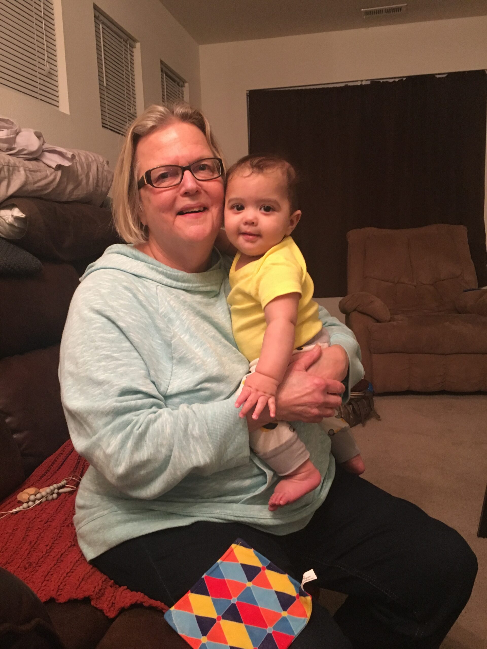 Woman with short blond hair and glasses sits on a couch, holding a smiling baby in a yellow shirt; a colorful geometric toy lies on a red rug nearby in a living room.